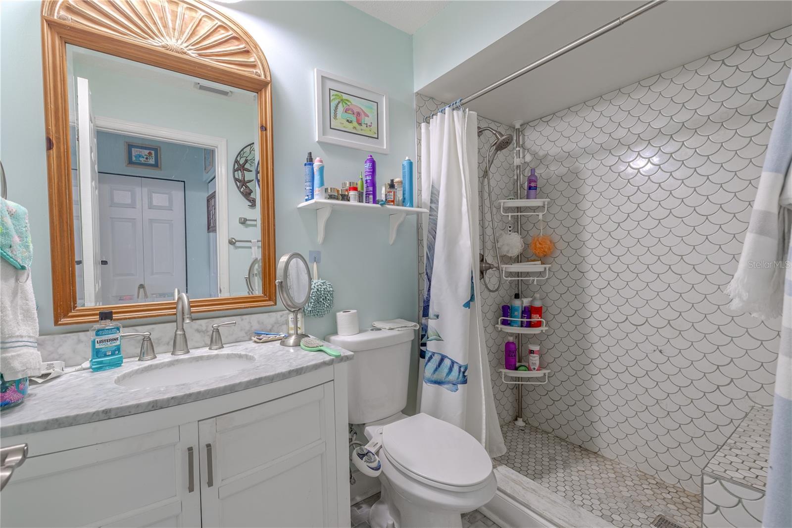 The en-suite primary bathroom combines style and comfort with thoughtful updates throughout. A white vanity with marble-look countertop provides plenty of counter space and storage, accented by brushed nickel fixtures and a large framed mirror. The walk-in shower features elegant mermaid-scale tile, a built-in bench, and a rainfall showerhead—bringing both charm and functionality to your daily routine. Soft lighting and a neutral color palette create a soothing atmosphere, making this space a true extension of the relaxing primary suite.