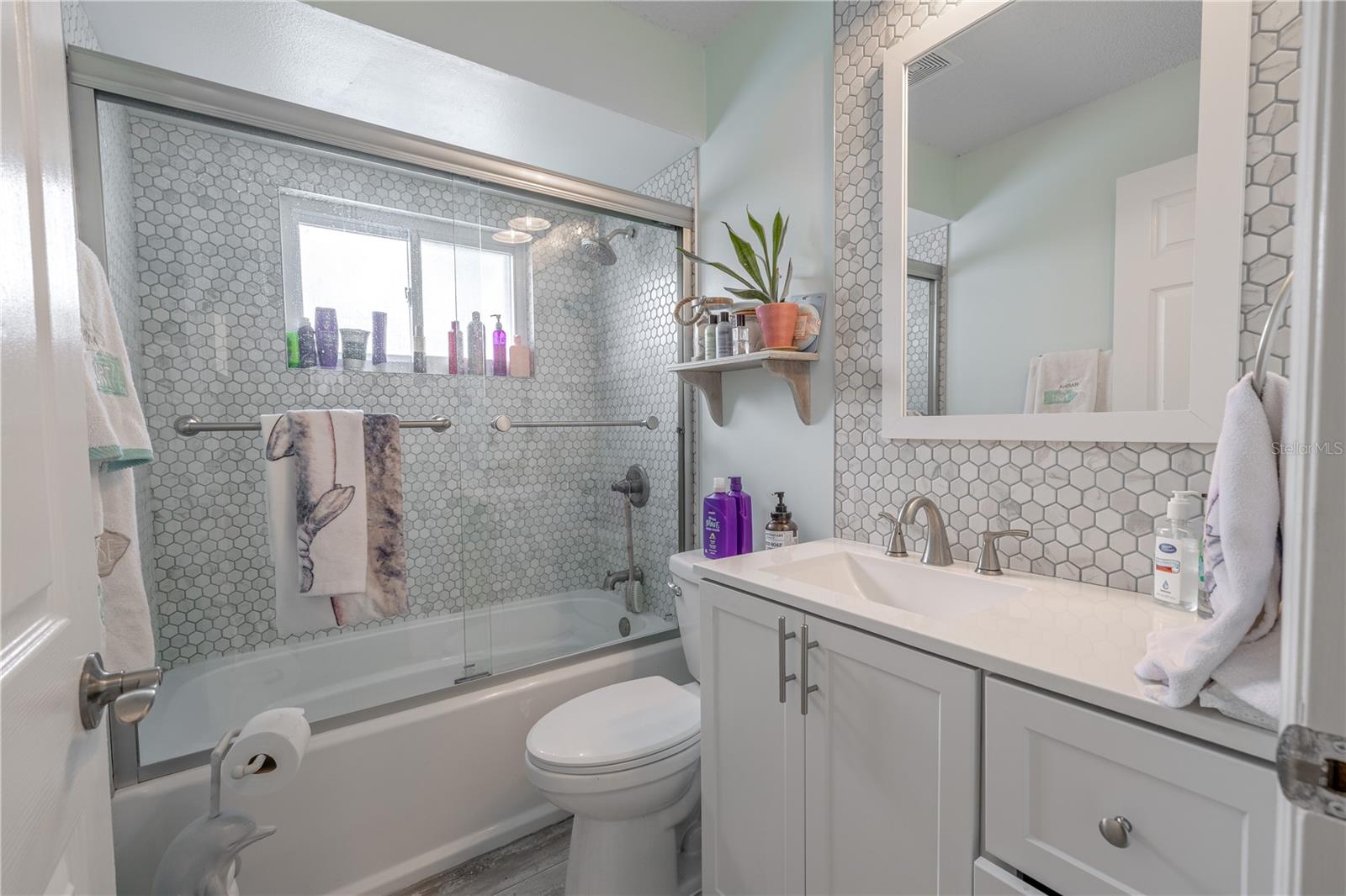 The second full bathroom features modern updates and stylish finishes throughout. A sleek vanity with quartz countertop and brushed nickel fixtures is paired with a framed mirror and hexagon-tiled accent wall for a touch of elegance. The tub and shower combo includes glass sliding doors and floor-to-ceiling tile in a coordinating hex pattern, creating a cohesive and contemporary look. A window above the shower brings in natural light, making the space feel bright and inviting—perfect for guests or shared household use.