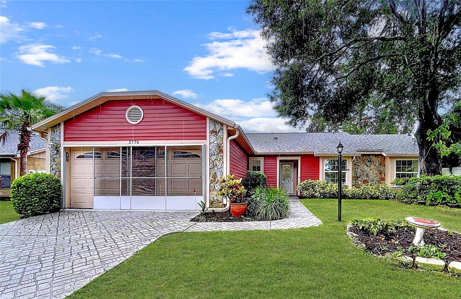 Paver-style driveway leads to a two-car garage with screened door enclosure and built-in cabinetry. Low-maintenance landscaping and potted plants add character to the front entry.