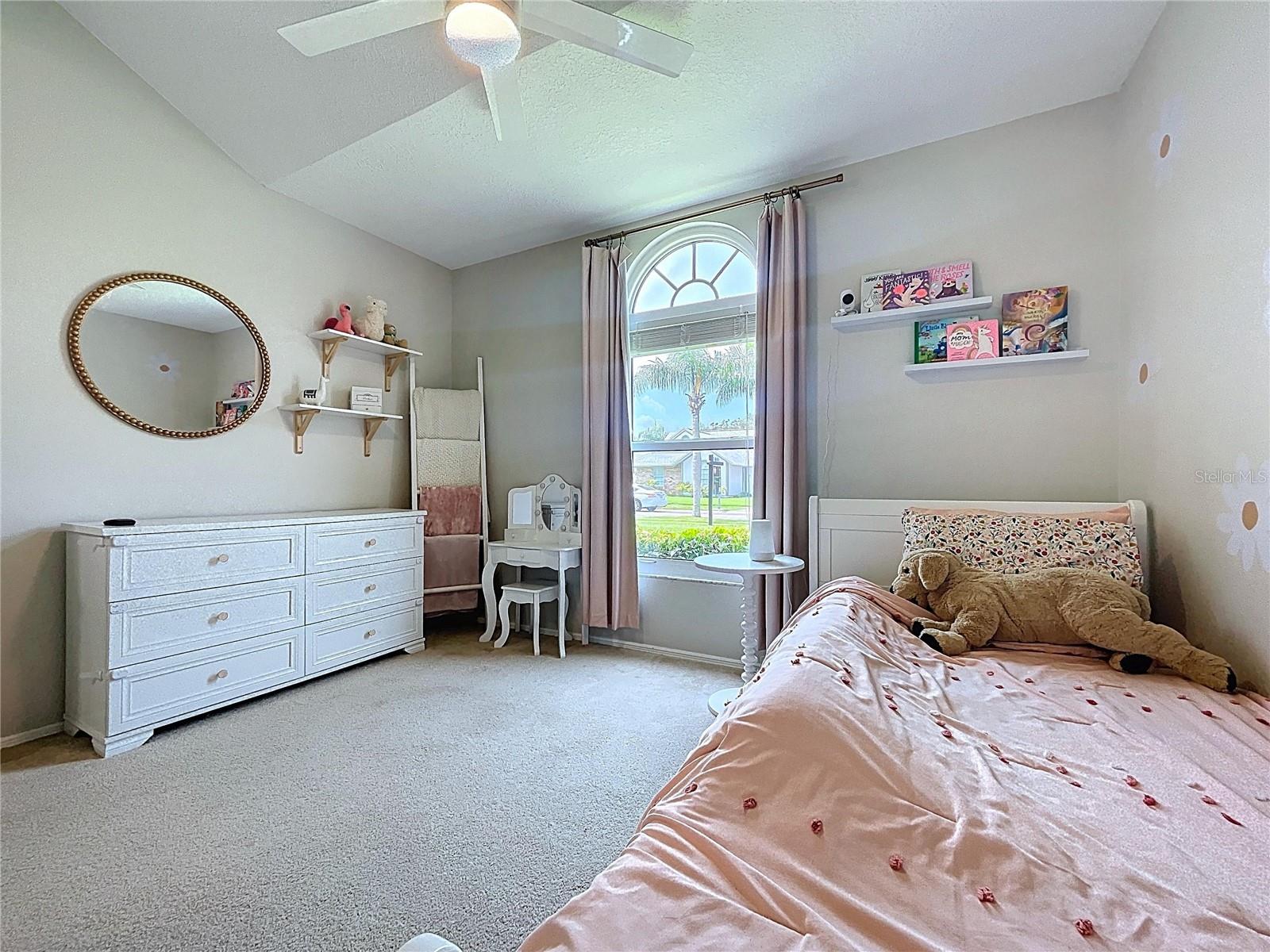 Bright and cheerful bedroom with soft carpeting, an arched window, and charming decorative details ideal for a child’s room or guest suite.