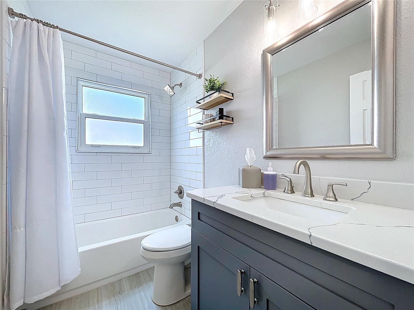 Stylishly updated bathroom featuring a subway-tiled shower/tub combo, floating shelves for added storage, and a contemporary navy vanity with a framed mirror.