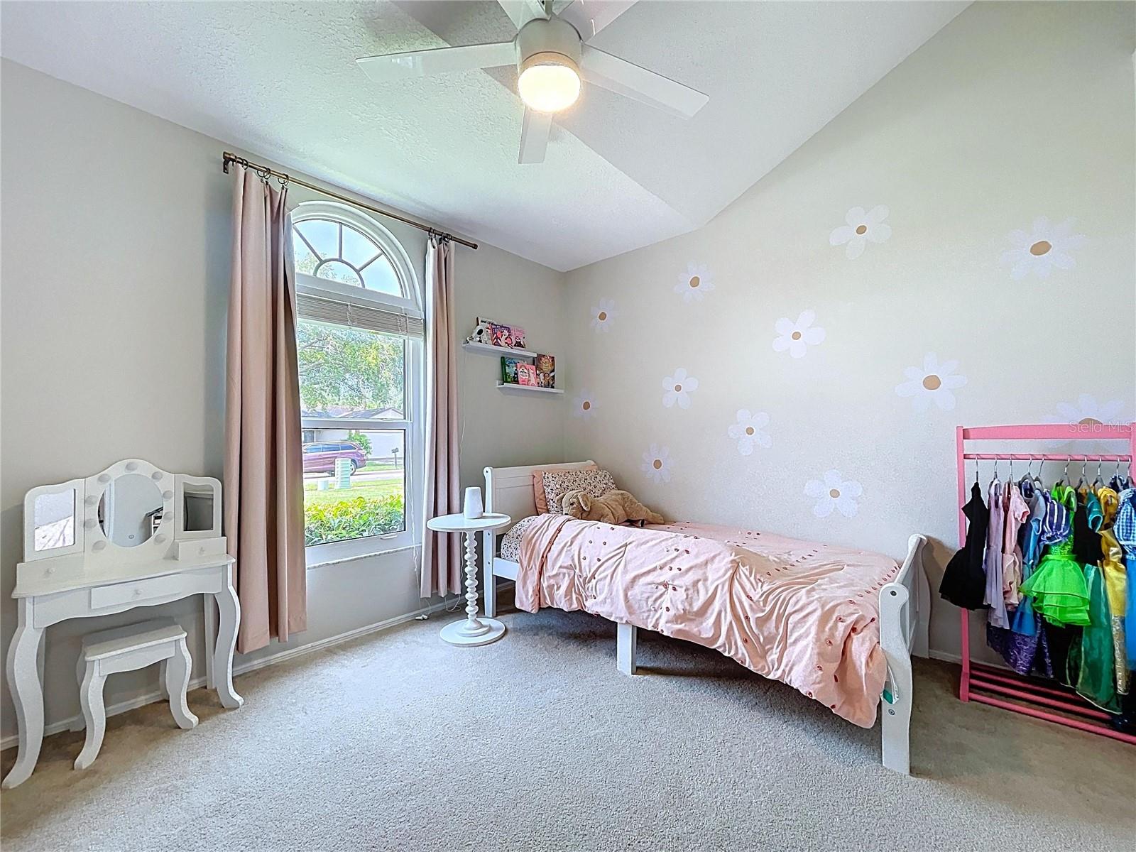 Light-filled bedroom with vaulted ceiling, charming arched window, and soft carpet. Custom painted daisy accents add a playful, cheerful touch.