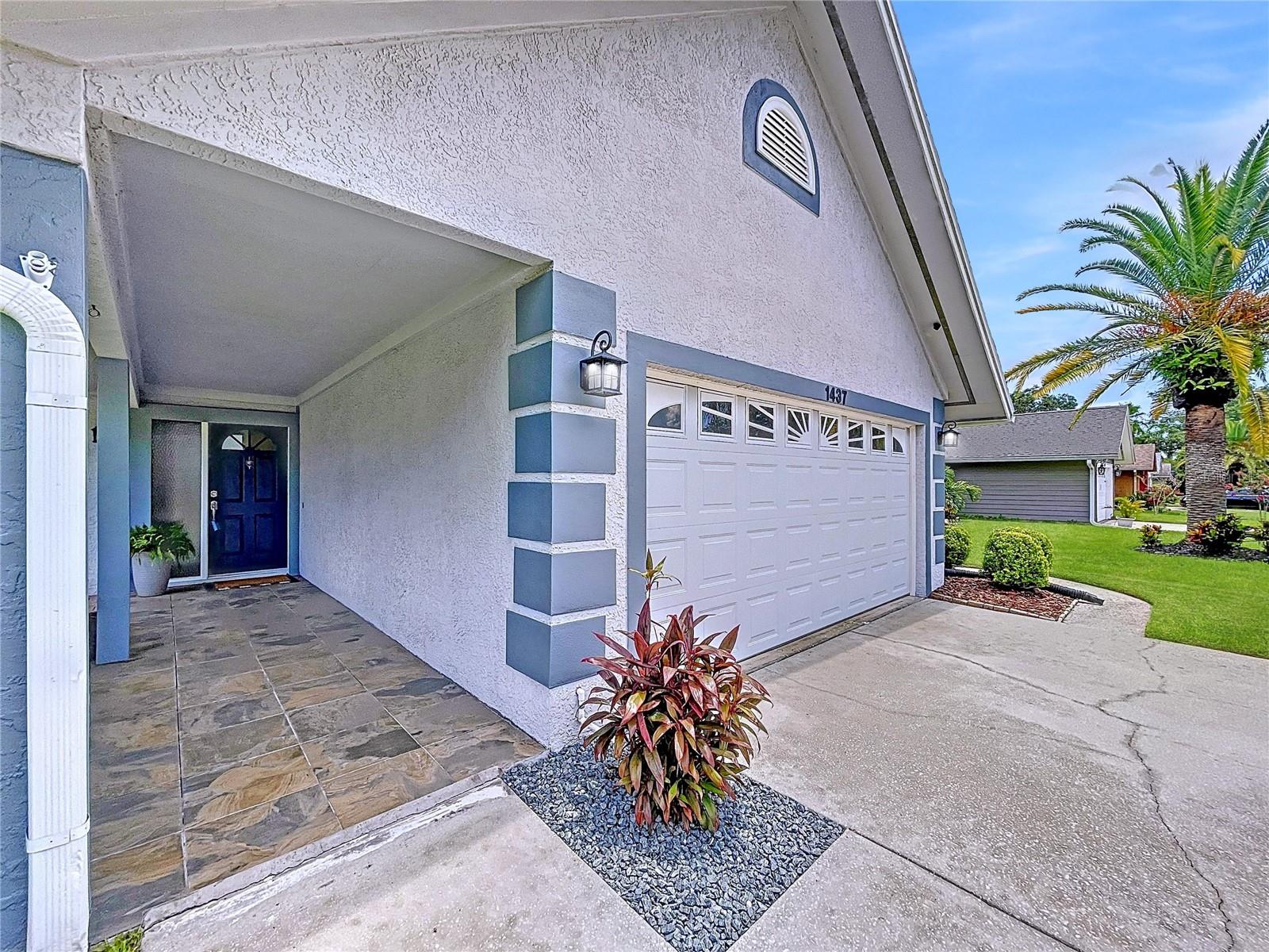 A welcoming covered entryway with slate-style tile leads to a charming front door and a two-car garage with decorative windows for added curb appeal.