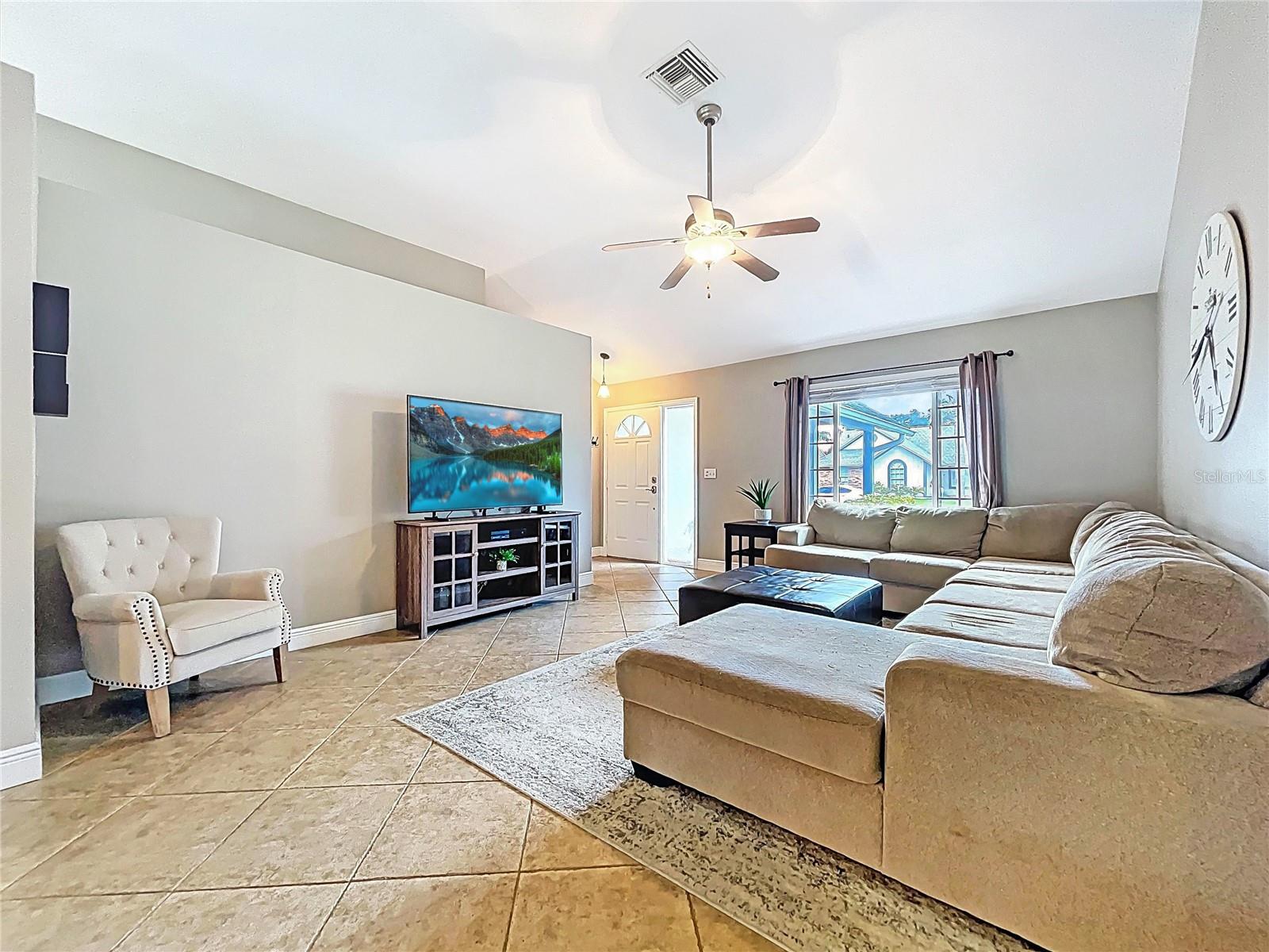 This airy living space features vaulted ceilings, oversized tile flooring, and a large picture window offering neighborhood views. A neutral palette complements the expansive sectional layout.