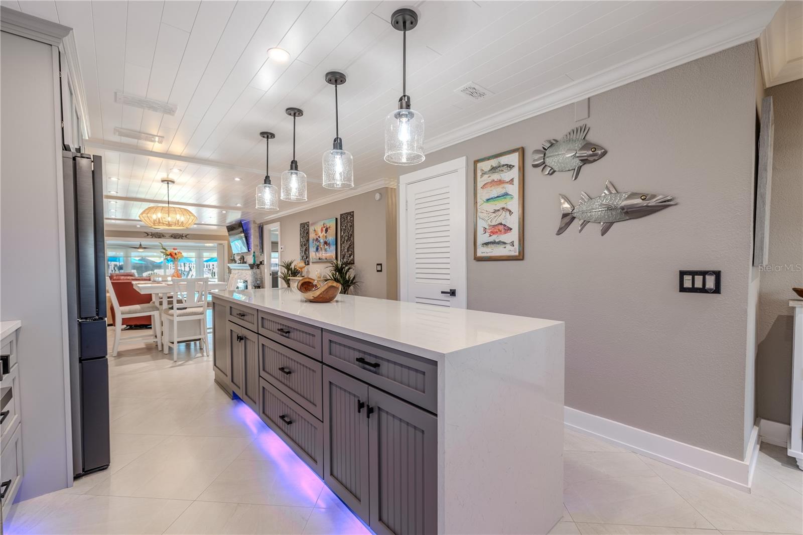 This striking kitchen island serves as both a visual centerpiece and a functional workhorse. Topped with a sleek, waterfall-edge quartz countertop, the island offers expansive prep space and seating for casual dining or entertaining. The base features custom cabinetry in a rich, muted gray with vertical panel detailing and matte black hardware, blending modern coastal style with timeless craftsmanship.
