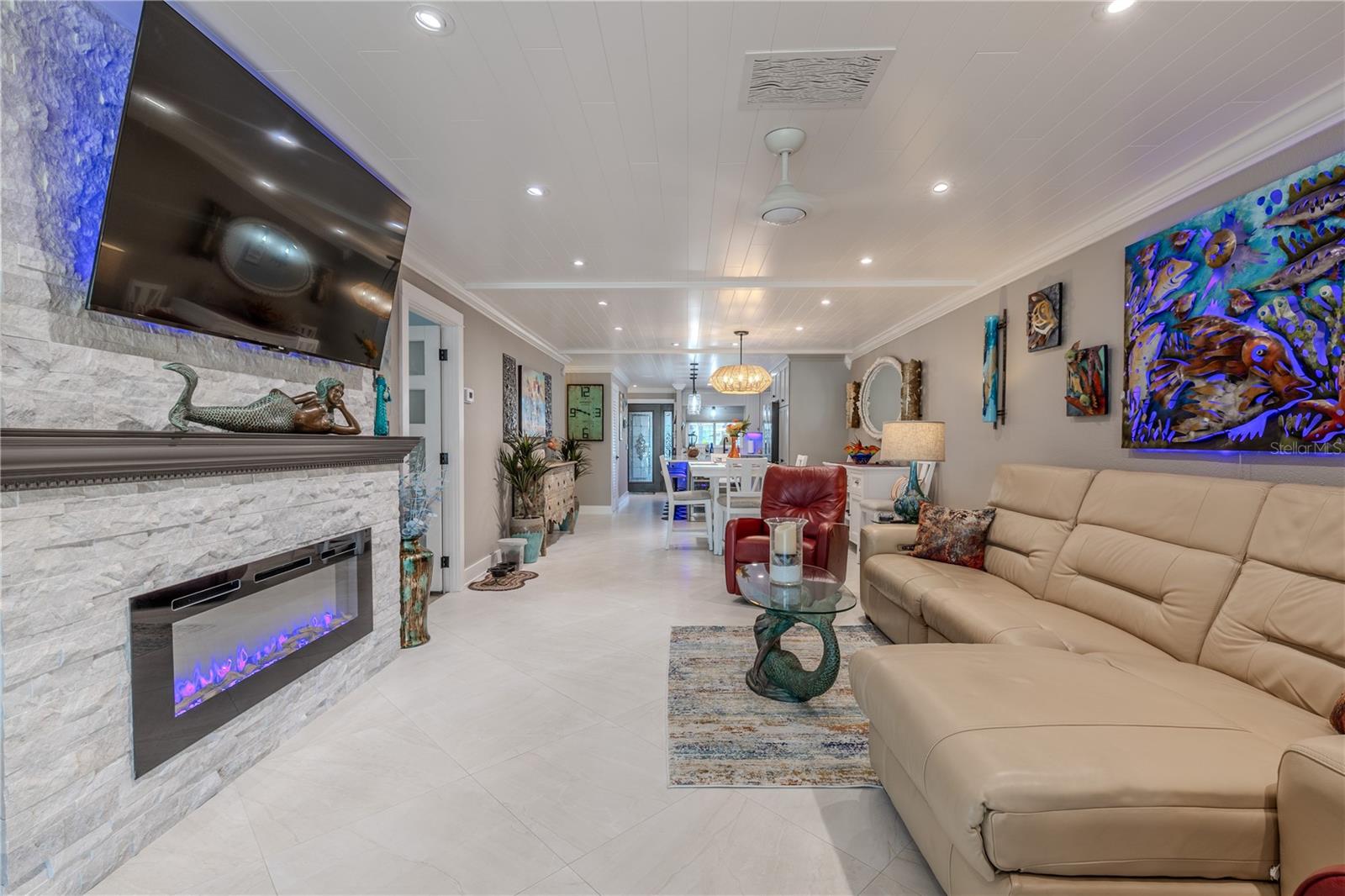 Beautifully designed open-concept living area featuring a custom white stacked stone fireplace with a built-in electric insert that offers both ambiance and supplemental heating. A WiFi-enabled thermostat allows for remote climate control, adding modern convenience. The space is enhanced by recessed LED lighting, crown molding, and a crisp white shiplap ceiling, creating a bright and polished coastal feel throughout.