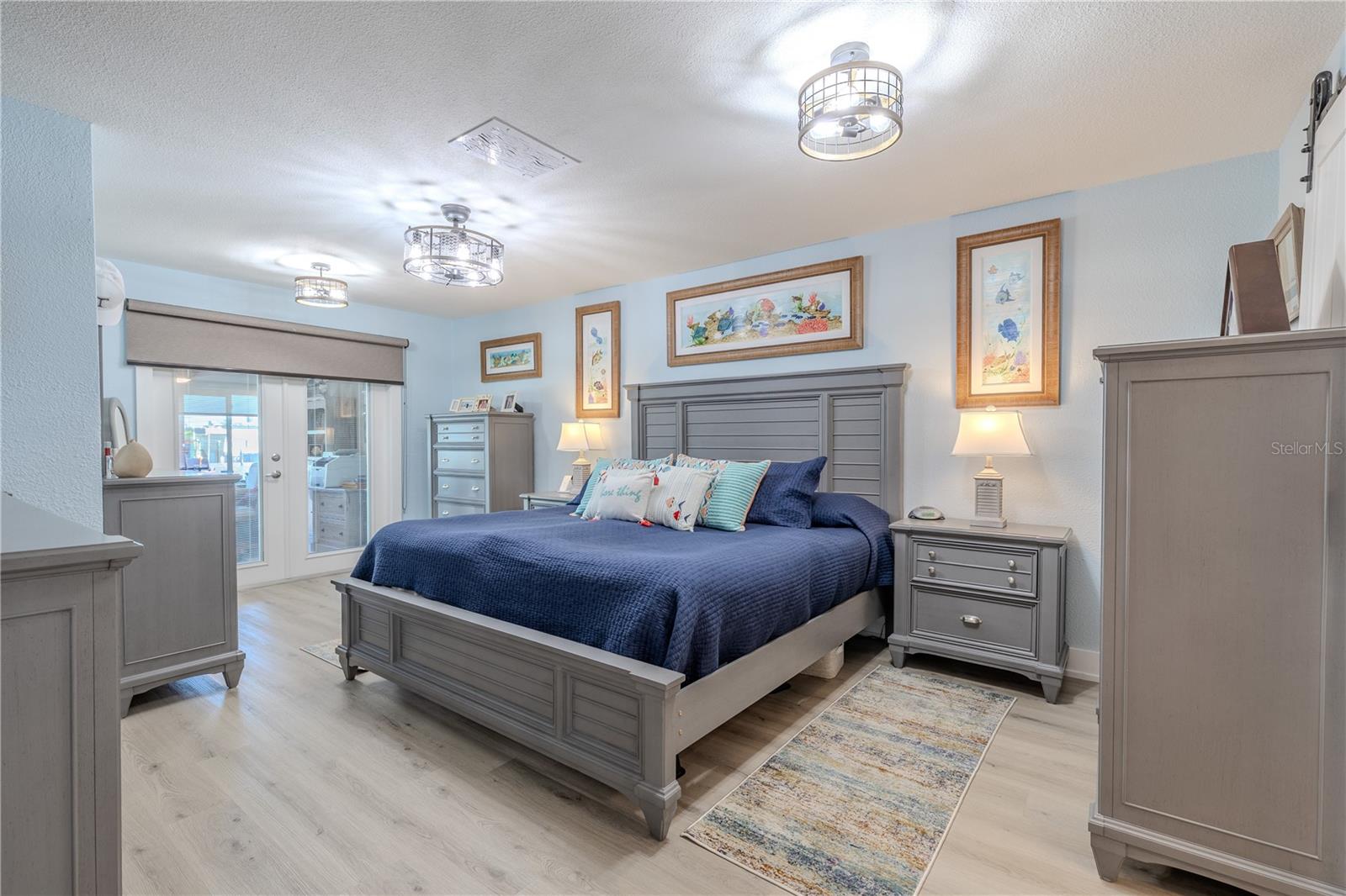 The primary bedroom combines comfort, function, and a bright, coastal vibe. Wide-plank luxury vinyl flooring and soft, neutral tones set the stage for effortless style, while modern caged light fixtures add a designer touch. Glass French doors open directly to the Florida room, allowing easy access to natural light, water views, and extended living space. This room checks all the boxes for everyday ease with just the right amount of wow.