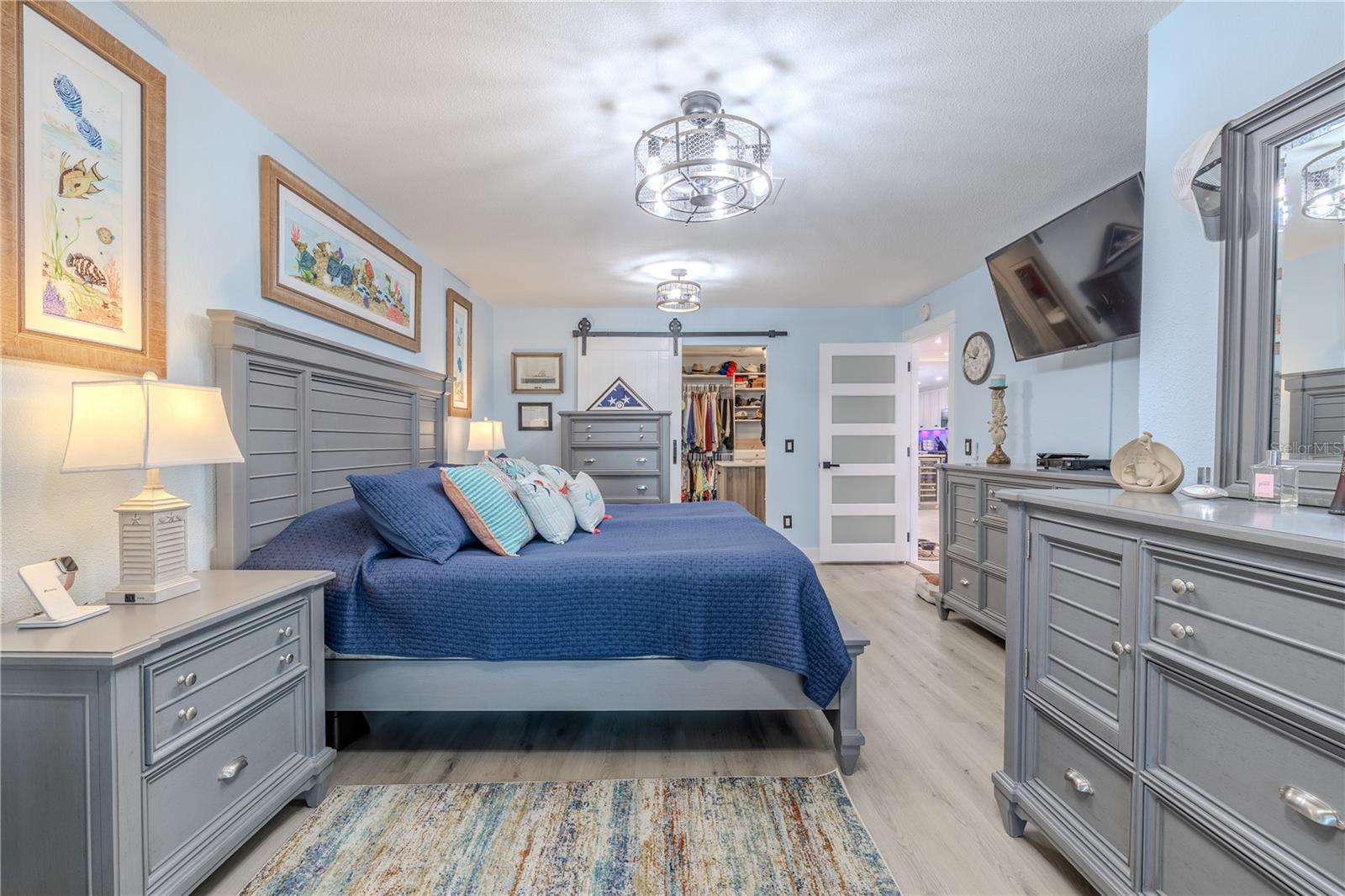 The primary bedroom blends comfort and functionality with crisp finishes and a bright, welcoming feel. Luxury vinyl plank flooring, modern caged ceiling fixtures, and soft coastal tones create a fresh, move-in ready space. A large walk-in closet with barn door access offers generous storage and organization.