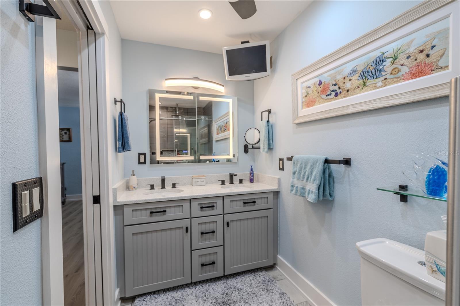 The updated primary bath blends modern function with clean, coastal style. A granite countertop with dual sinks sits atop new custom cabinetry featuring soft-close hinges. The built-in LED-lit medicine cabinet offers adjustable lighting, inside and out, with a built-in defogger for added convenience. Four recessed LED can lights brighten the space, while a ceiling fan with light and a mounted TV add comfort and functionality. Every detail is designed for ease, efficiency, and everyday luxury.