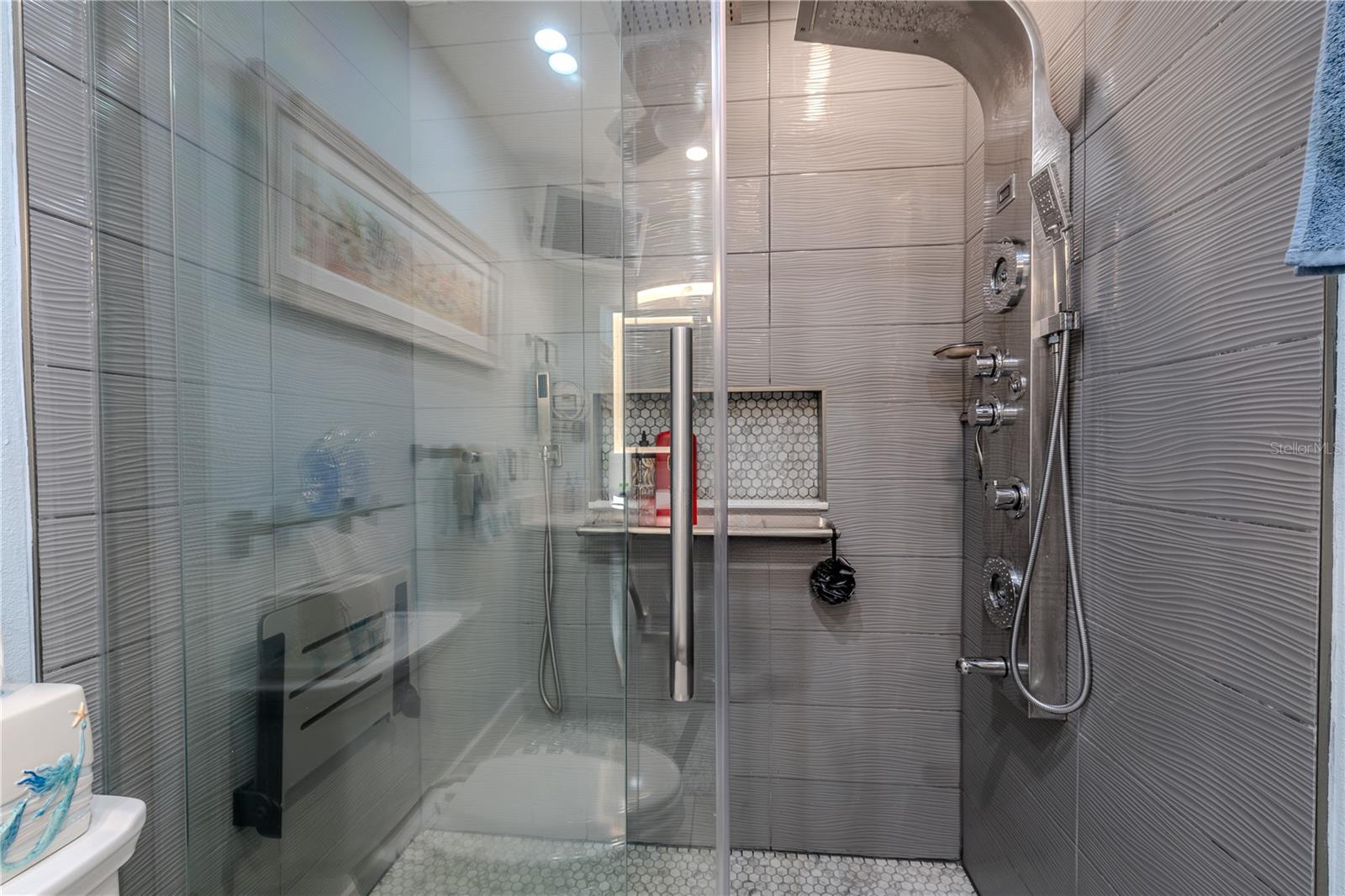 The oversized 36" x 58" low-threshold shower is enclosed in sleek glass doors and designed with both luxury and function in mind. A 4-foot wall-mounted shower panel offers multiple jets, a rainfall head, and handheld wand, while a separate sprayer and control near the fold-down bench add comfort, safety, and accessibility. Textured tile walls and a tiled niche complete this thoughtfully designed space.