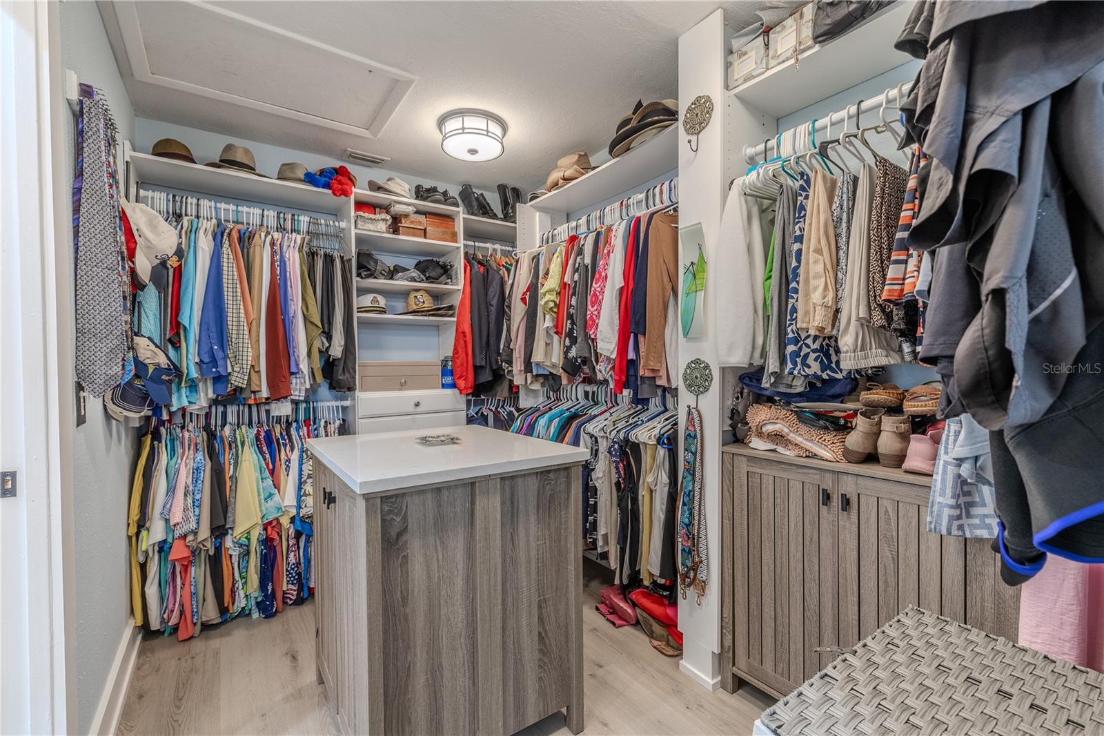Primary Closet: A center island with quartz countertop provides additional storage and folding space, while upper display shelves keep seasonal items or accessories neatly tucked away. Luxury vinyl plank flooring and overhead lighting complete the clean, tailored feel of this well-appointed storage space.