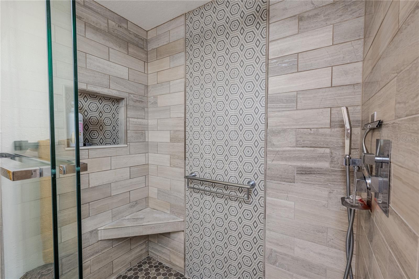 The walk-in shower spans approximately 36" x 59" and features sleek glass sliding doors, a rainfall shower head, corner seat, and built-in niche shelving. A bold geometric tile accent wall adds modern texture, while the ultra-quiet exhaust fan—with its own control—keeps the space fresh without the hum.