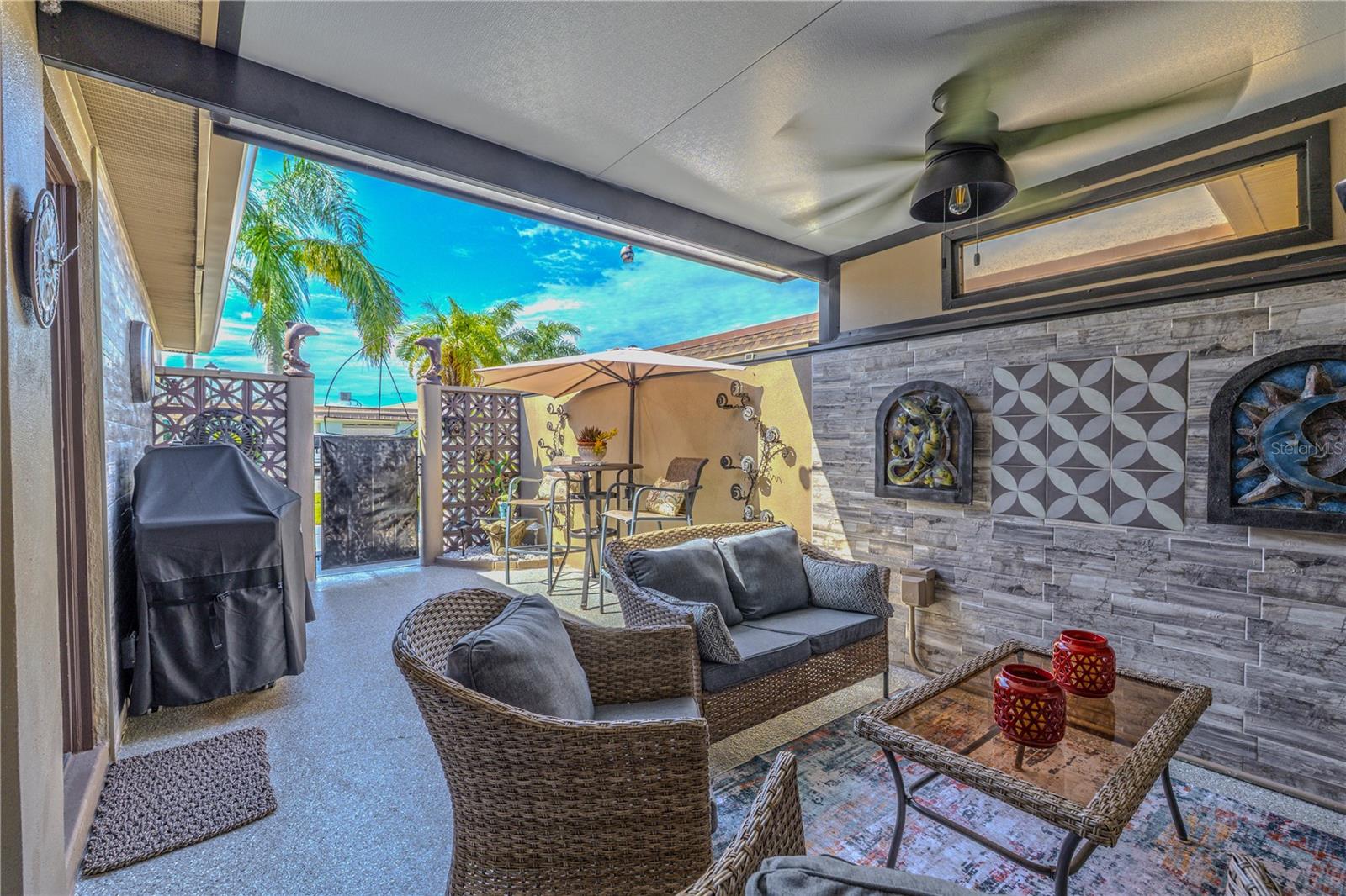 A welcoming covered front lanai/patio brings the outside-in with a transom window, new epoxy surface floor with a lifetime warranty, two custom tile feature walls, sitting area, TV, and ceiling fan. private, resort-style lanai that brings the indoors out. The covered section offers a shaded seating area with modern ceiling fan, textured tile accent wall, and space to relax or entertain in comfort. Just beyond, an open-air patio is perfect for alfresco dining, ideal for a bistro table and umbrella. Surrounded by mid-century inspired breeze block privacy walls, this outdoor oasis feels like your own hidden escape—just steps from home.