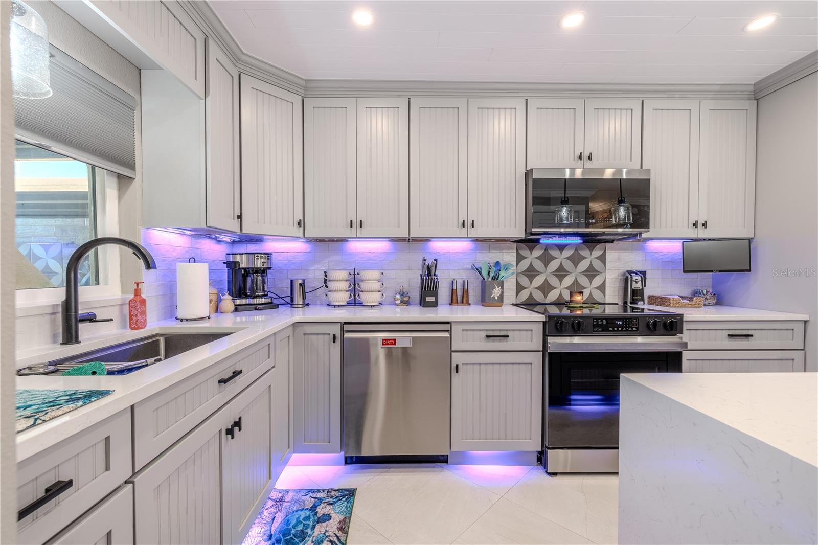 The L-shaped gourmet kitchen  seamlessly blends coastal charm with sleek, modern updates. Soft beadboard cabinetry with matte black hardware creates a clean, tailored look while providing ample storage from floor to ceiling. The bright quartz countertops offer generous workspace and are beautifully complemented by a subway tile backsplash that adds depth and texture. Beneath the upper cabinets, custom LED lighting in a soft violet-blue hue creates ambient illumination and a contemporary vibe, echoed by matching toe-kick lighting below the base cabinets.