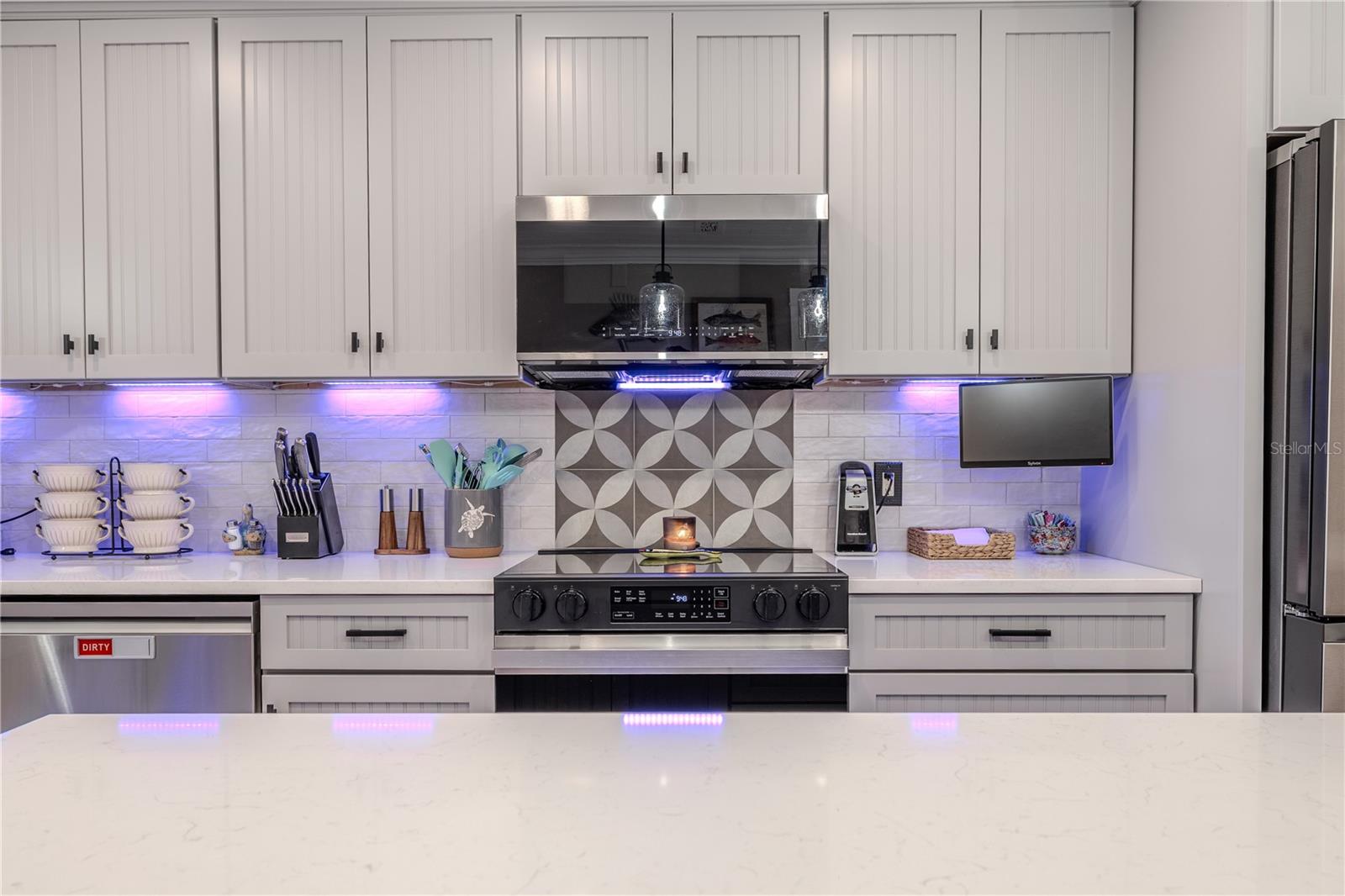 This beautifully framed view of the kitchen highlights the home’s impeccable attention to detail and modern coastal style. Centered on the back wall, a bold patterned tile backsplash behind the sleek black Samsung electric range creates a dramatic focal point, framed by clean, white quartz countertops and soft gray shaker-style cabinetry with beadboard detail and matte black hardware.