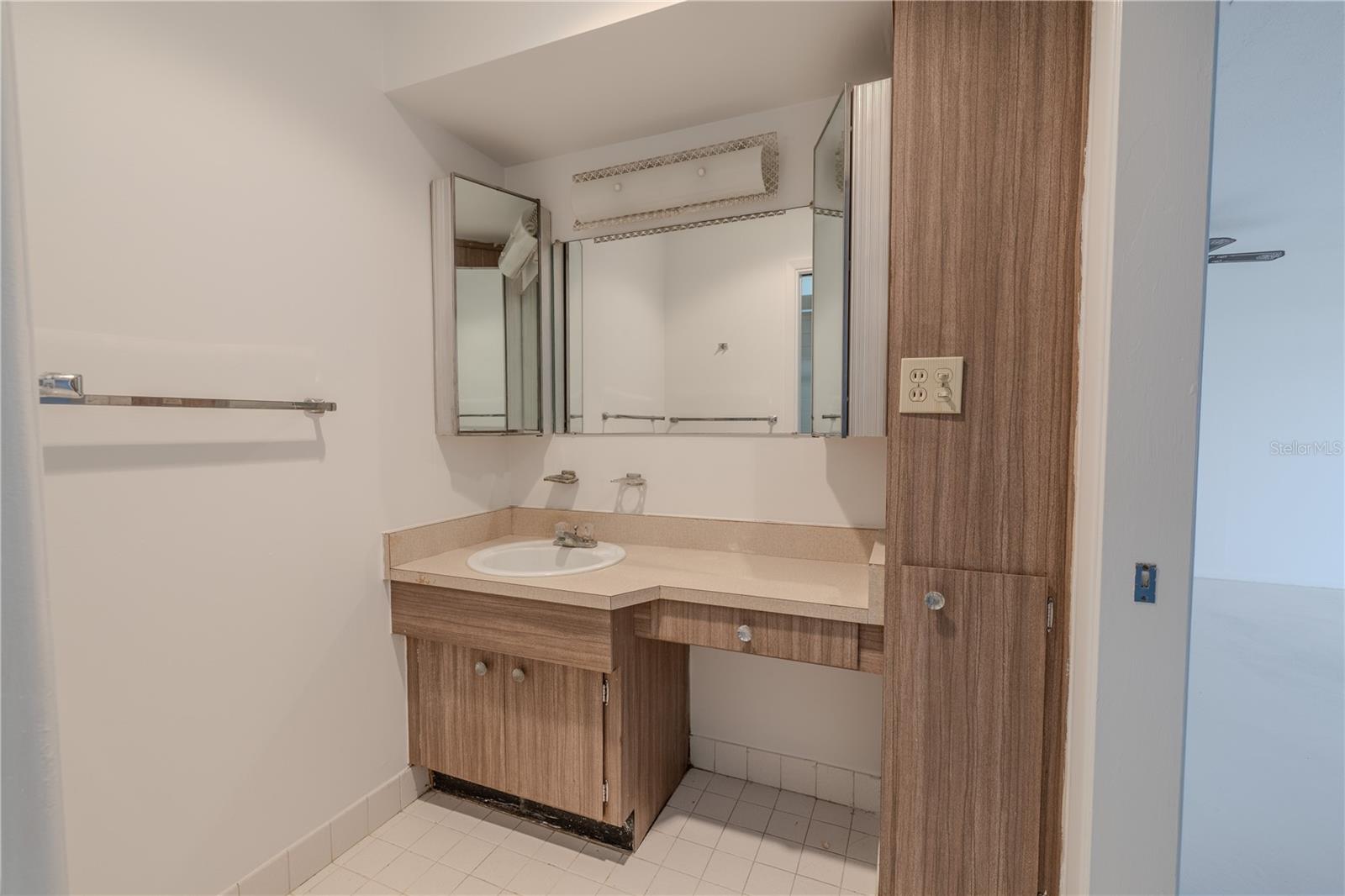 The bathroom features a practical and spacious layout with a retro flair. A long, angled vanity provides ample counter space and storage, topped with a built-in sink and surrounded by wood-toned cabinetry and mirrored medicine cabinets for added functionality. The tile flooring and neutral tone walls keep the space feeling clean and bright.