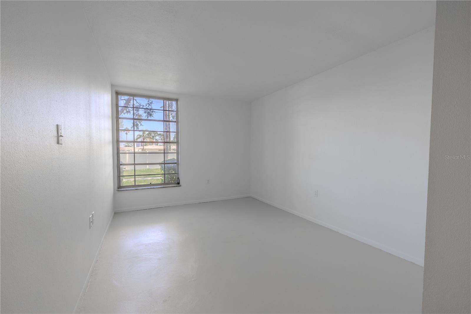 The primary bedroom offers a peaceful, sunlit retreat with a large picture window that frames serene views of the manicured courtyard and allows natural light to flood the space. The freshly painted white walls and matching floors create a clean, calming atmosphere with a minimalist feel—perfect for rest and relaxation. Generously sized and thoughtfully laid out, the room provides plenty of space for a queen or king-size bed along with additional furnishings. Whether you keep it simple or add your personal touch, this bright and airy room is a blank canvas ready to become your private sanctuary.