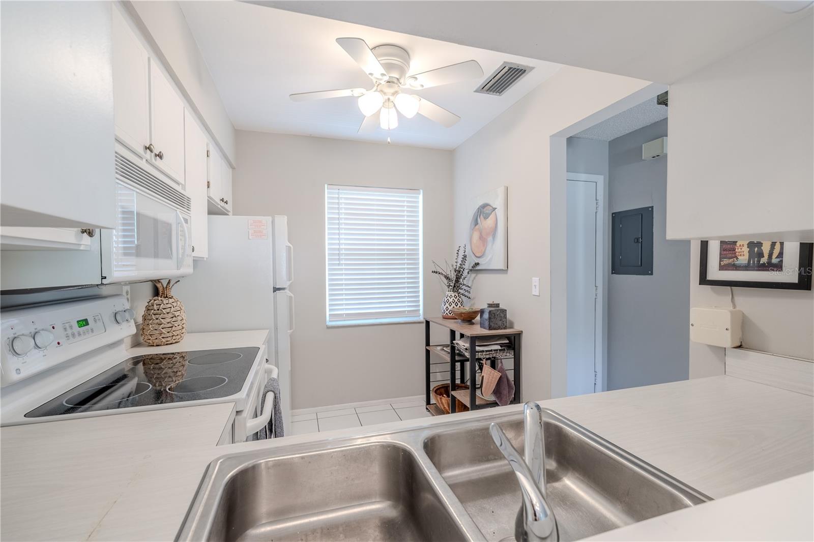 This light and bright kitchen features a cozy dining nook with a large window that fills the space with natural light. A ceiling fan overhead adds comfort, while the neutral paint palette and crisp white cabinetry create a clean, inviting atmosphere. The functional layout includes a double-basin stainless steel sink with a pass-through opening to the living area.