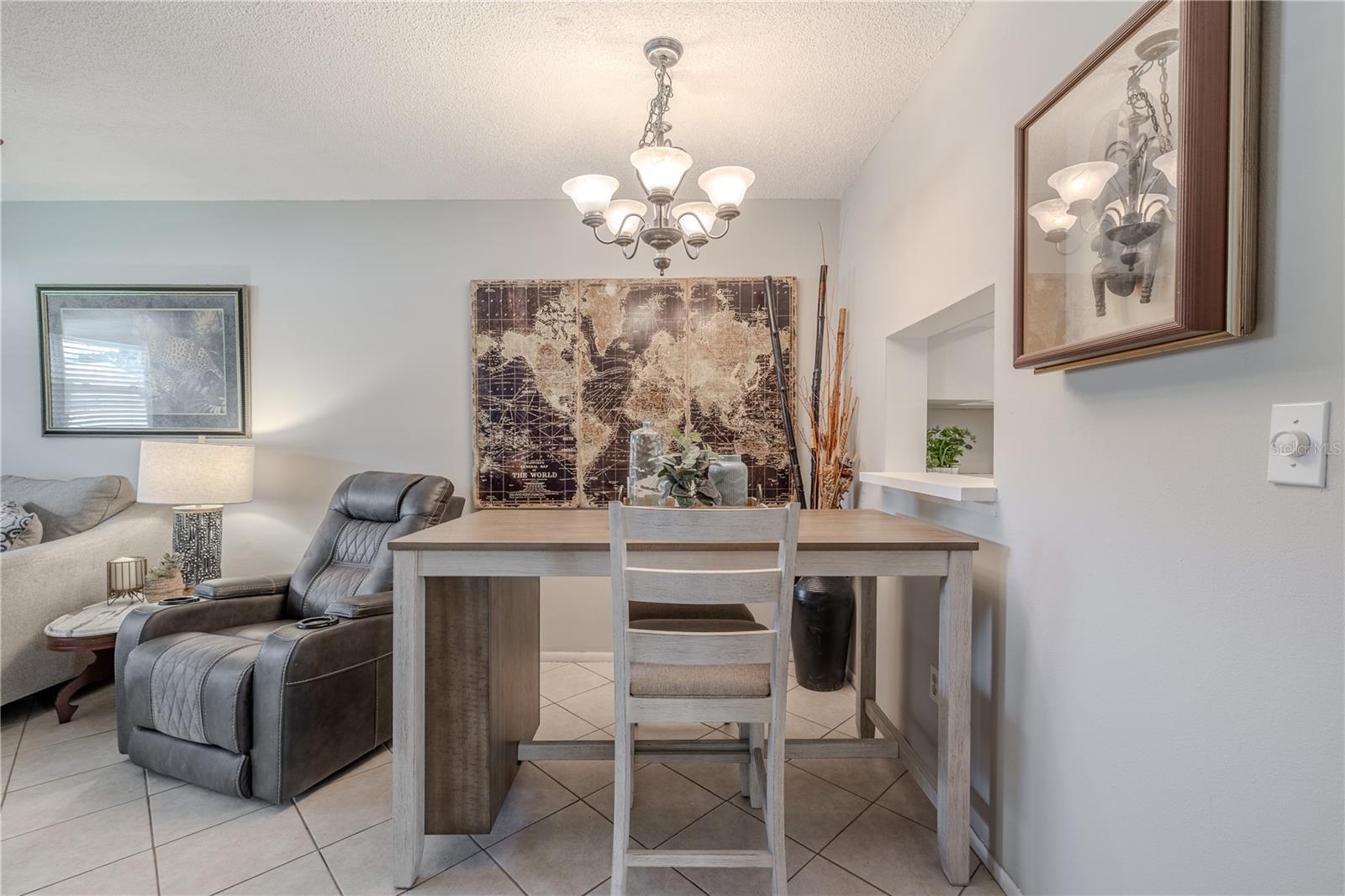 The dining area offers a cozy and versatile space with neutral tones, tile flooring, and a stylish chandelier that adds a touch of elegance.The convenient pass-through window to the kitchen makes serving and entertaining a breeze. Perfectly positioned between the living room and kitchen, this space can easily function as a casual dining nook or a work-from-home setup.