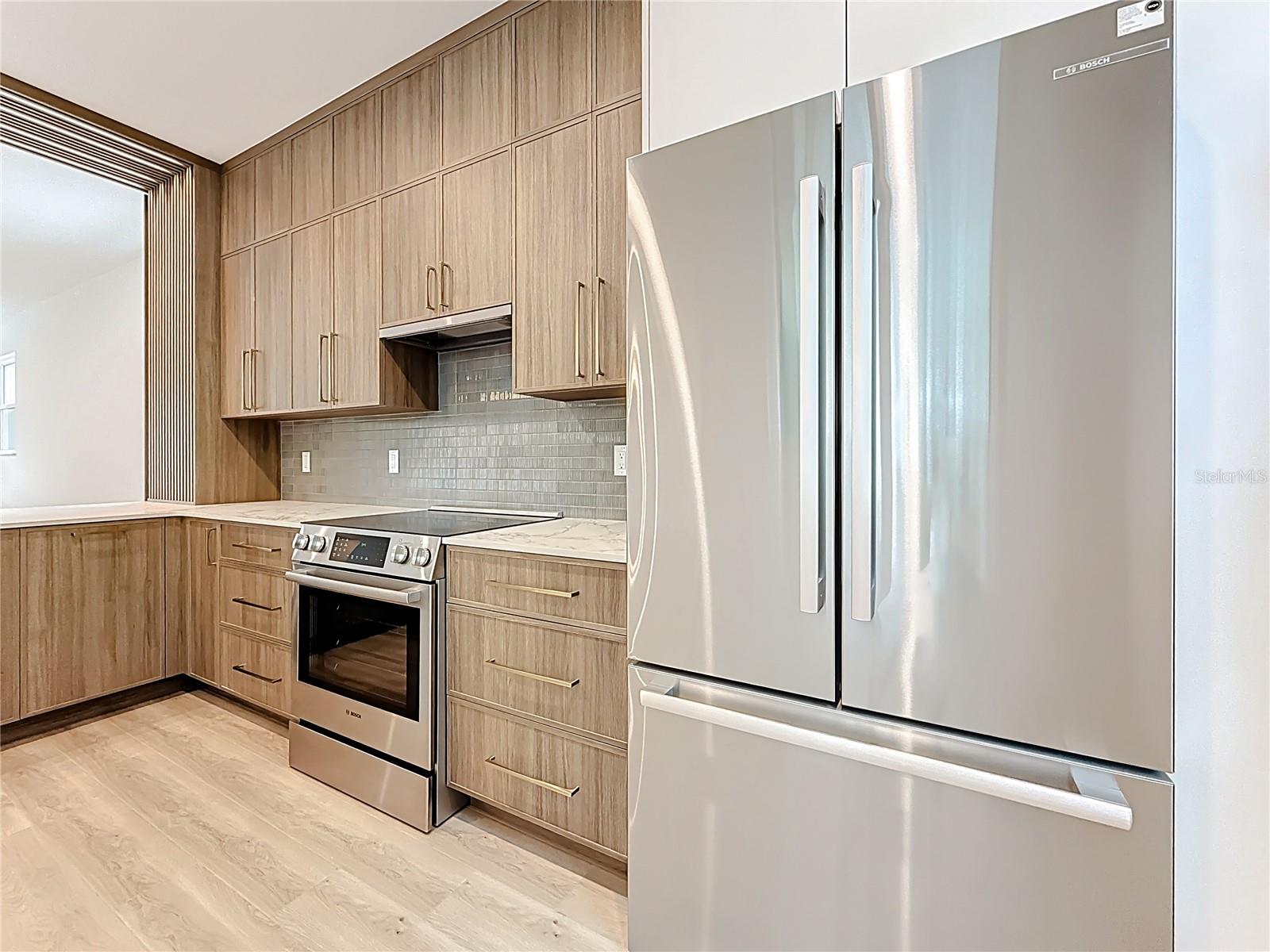 Sleek and modern kitchen featuring custom wood cabinetry, quartz waterfall countertops, stainless steel appliances, and stylish accent details — all bathed in natural light.