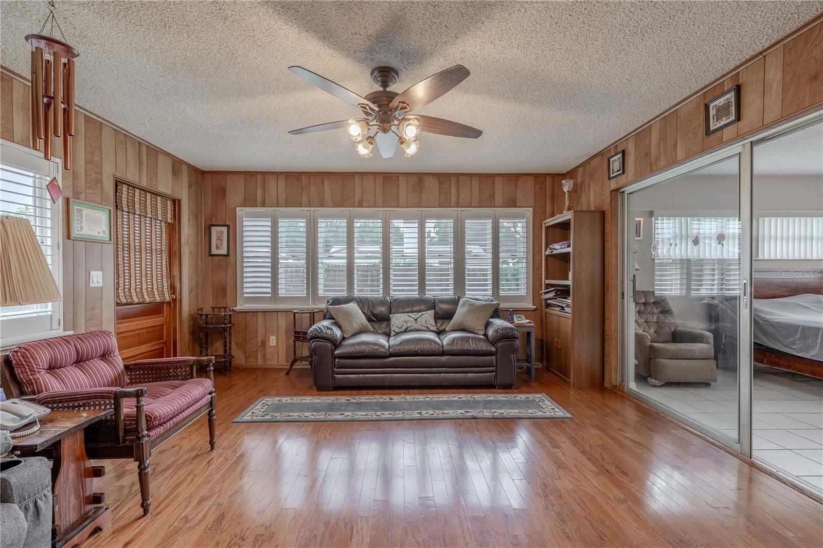 Light-filled Florida room with expansive windows fitted with plantation shutters, offering views of the backyard while allowing for adjustable natural light. Wood-look flooring adds warmth to the space, complemented by wood-paneled walls and a ceiling fan for year-round comfort. Sliding glass doors connect the room to the main living area, making it an ideal spot for relaxing, entertaining, or use as a flexible bonus space.