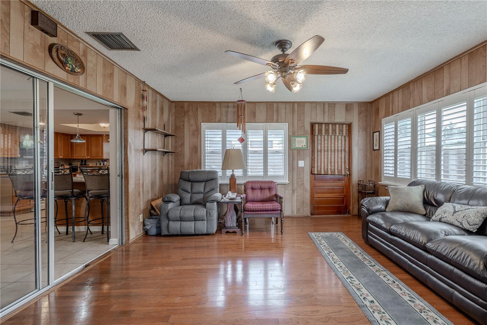 nviting Florida room with wood-look flooring, wood-paneled walls, and a ceiling fan for added comfort. Plantation shutters on multiple windows provide both privacy and natural light control. Sliding glass doors open to the kitchen and breakfast bar, while a solid wood exterior door offers direct backyard access, making this a versatile space for relaxation or entertaining.