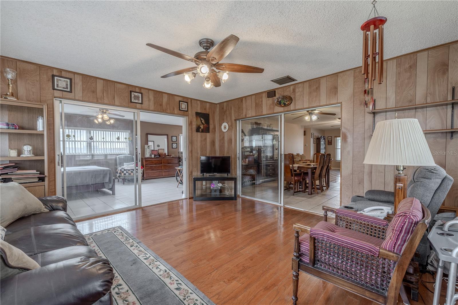 Spacious Florida room with wood-look flooring and warm wood-paneled walls, offering flexible living space with direct access to both the dining area and a bedroom via sliding glass doors. A ceiling fan provides comfort, while multiple access points and open sightlines make this room ideal for relaxing, entertaining, or creating a multi-purpose bonus space.