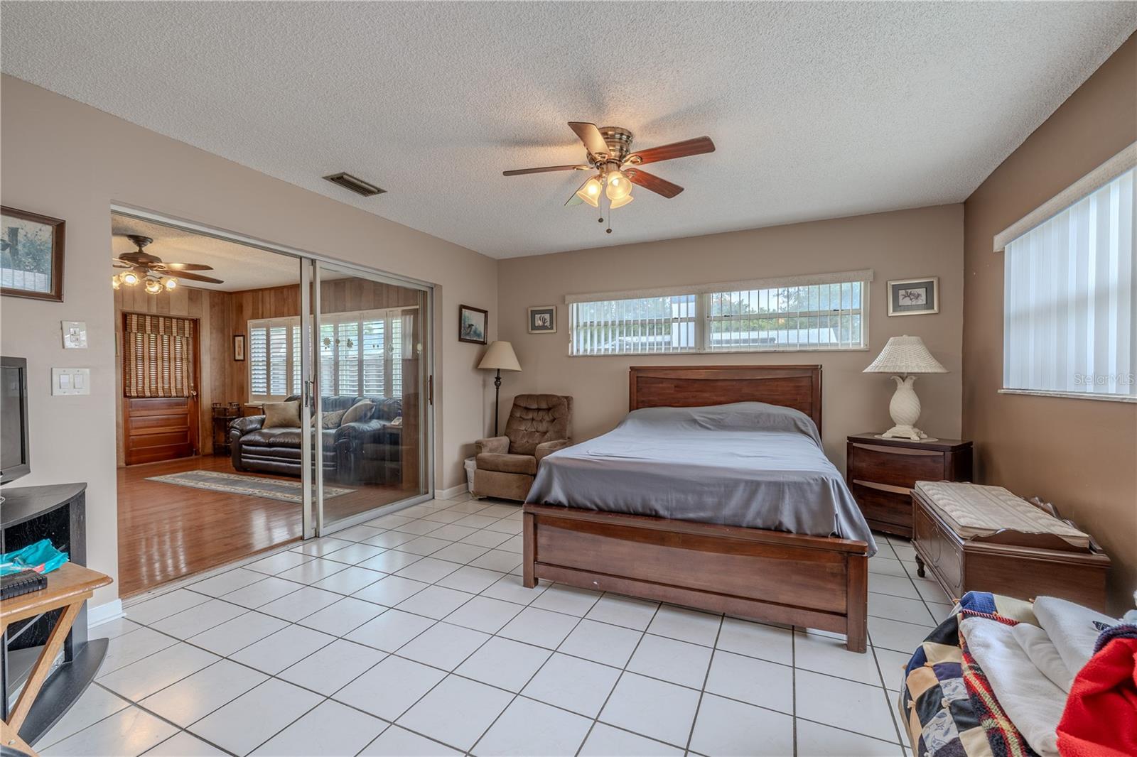 Bright primary bedroom with tile flooring, dual ceiling fans, and large windows that let in abundant natural light. Sliding glass doors provide direct access to the Florida room