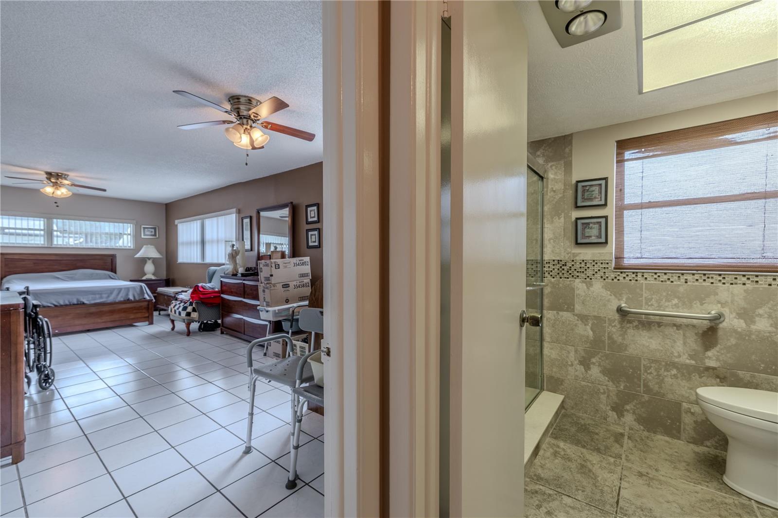 The spacious primary bedroom features tile flooring, dual ceiling fans, and abundant natural light from multiple windows. Just steps away, the updated en-suite bathroom offers a walk-in shower with decorative tile accents, a grab bar for accessibility, and a modern vanity. The bathroom includes dual entry doors—one from the bedroom and one from the hallway—for added convenience.