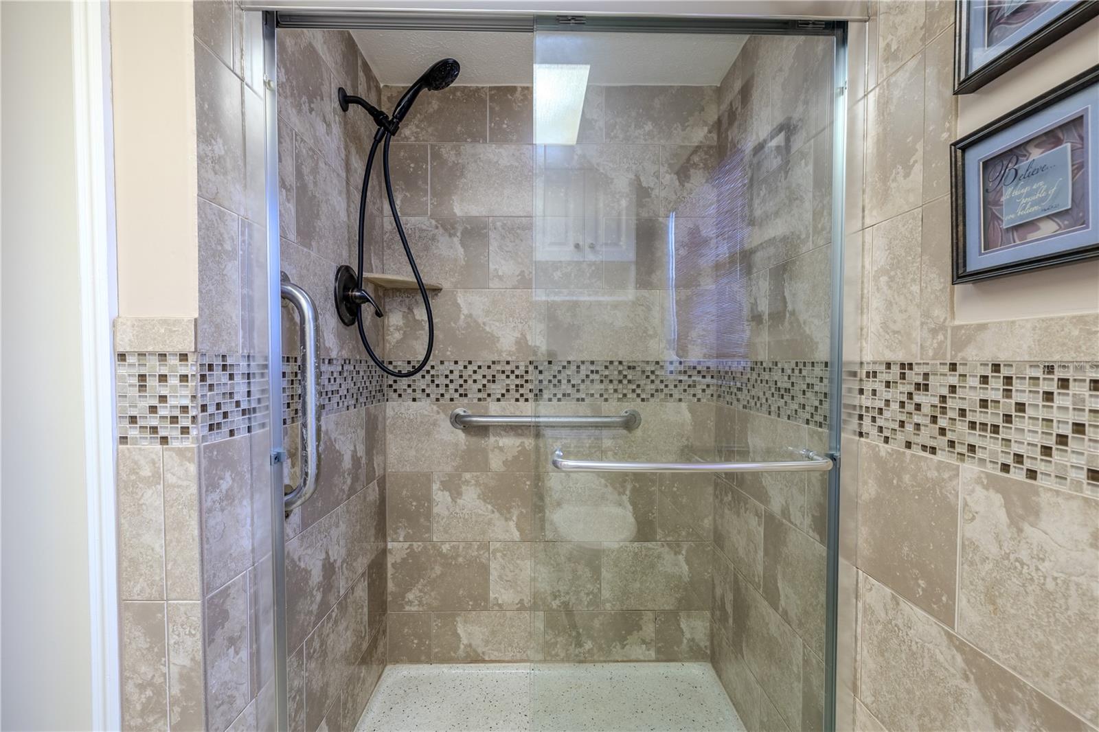 Beautifully tiled walk-in shower featuring a mosaic accent border, sliding glass doors, and built-in grab bars for safety. A handheld showerhead and built-in corner shelf add convenience, while the neutral tile design creates a modern, spa-like feel.