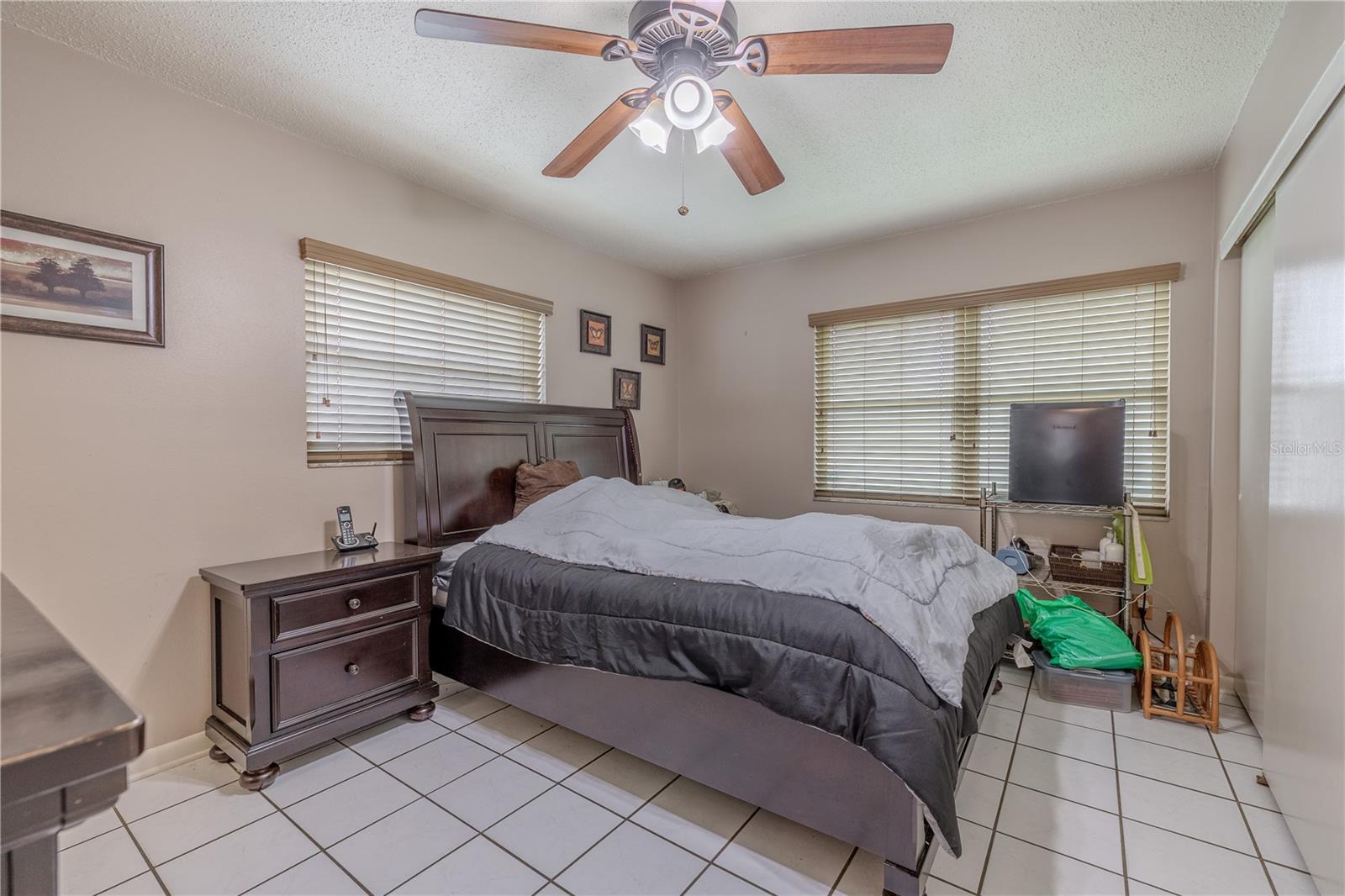 Bright second bedroom with tile flooring, neutral wall tones, and a ceiling fan for comfort. Large windows on two walls fill the space with natural light, and a double closet provides ample storage.
