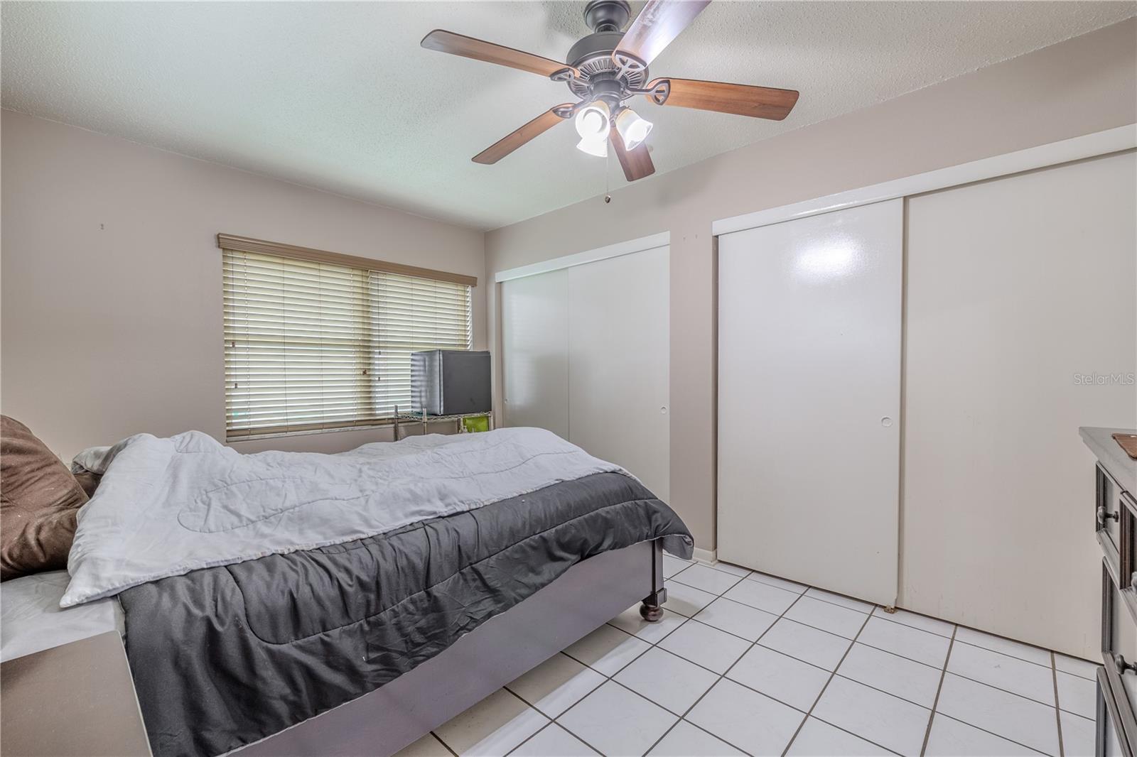Second bedroom features tile flooring, a ceiling fan, and a large window that brings in natural light. A wall-to-wall double closet provides generous storage space, making the room both functional and comfortable.