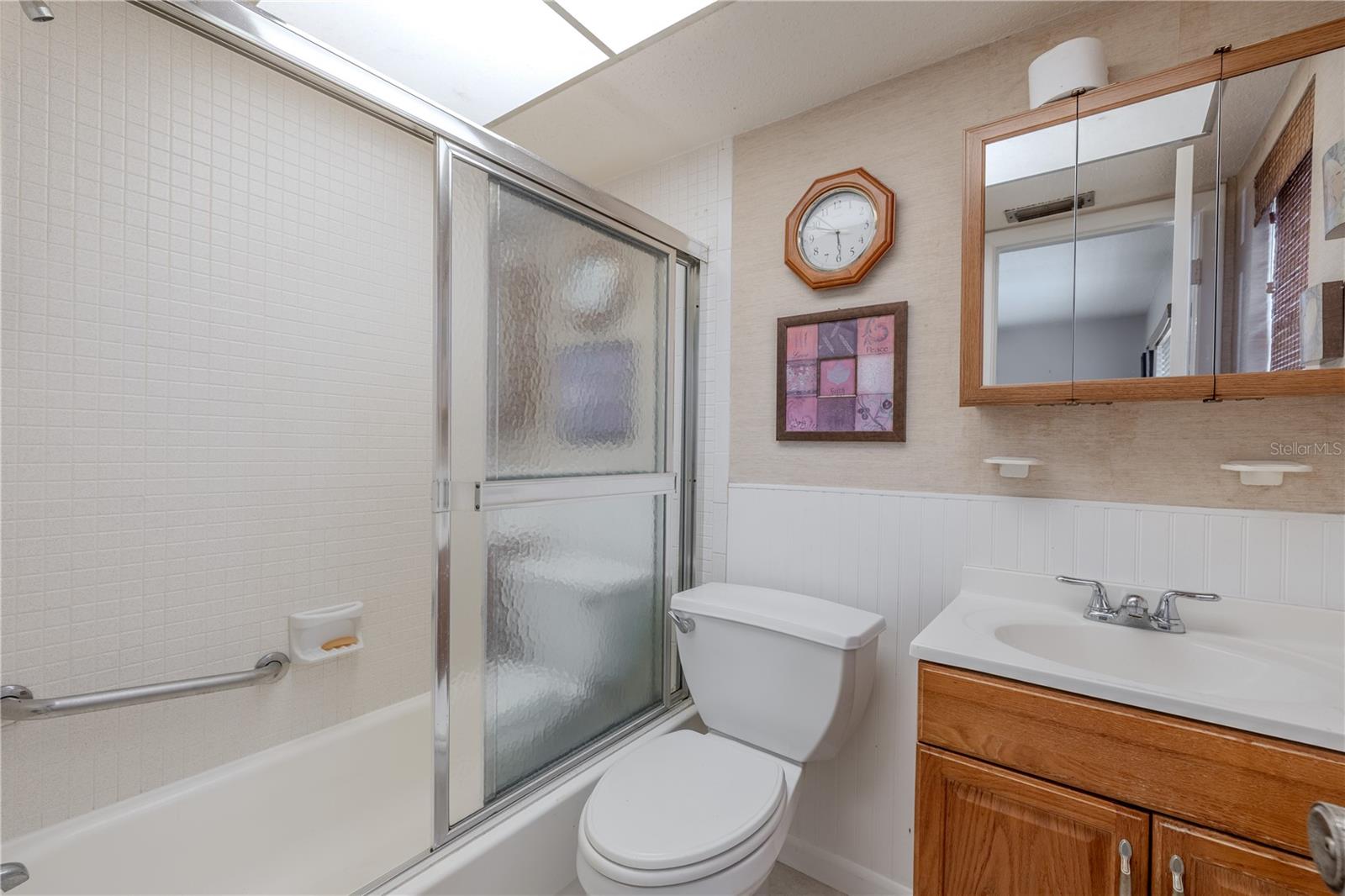 Full second bathroom featuring a tub/shower combo with sliding glass doors and a grab bar for added safety. Includes a vanity with storage, mirrored medicine cabinet, and overhead lighting. Neutral finishes and a large overhead light create a bright and functional space.