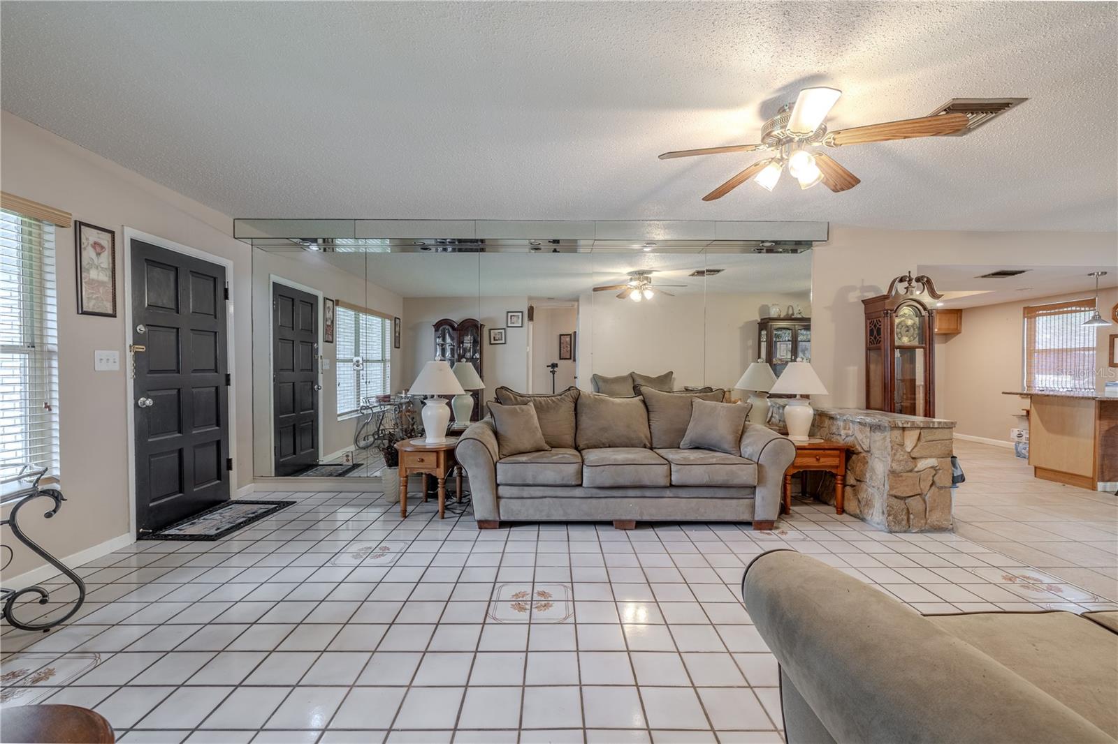 Inviting living room with tile flooring featuring decorative inlays, mirrored accent wall for enhanced light and space, and a ceiling fan for year-round comfort. The front entry opens directly into the living area, creating a welcoming flow, while large windows fill the space with natural light.