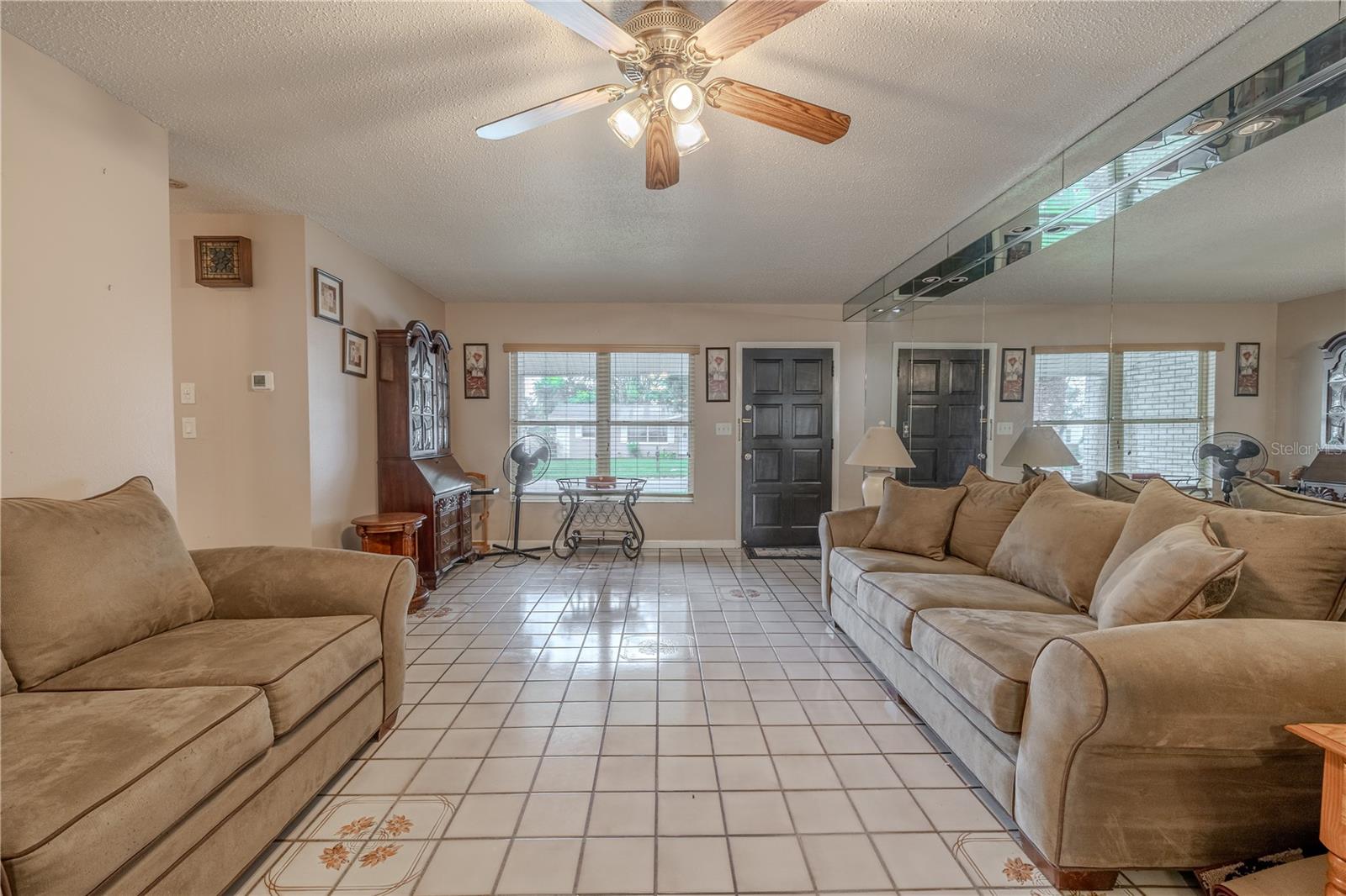 Bright and welcoming living room with large front-facing windows that fill the space with natural light. Neutral tile flooring with decorative accents runs throughout, complemented by a mirrored feature wall that enhances the sense of space. The entryway flows seamlessly into the seating area, where a ceiling fan adds comfort and style.