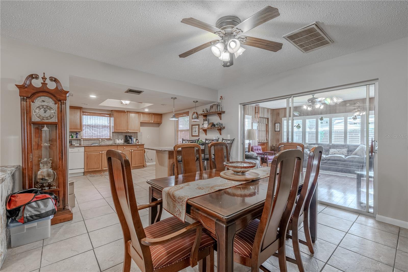 Spacious dining area seamlessly connected to the kitchen, featuring neutral tile flooring and a ceiling fan for added comfort. A wide opening to the kitchen allows for easy serving and conversation, while sliding glass doors lead to a bright sunroom, filling the space with natural light.