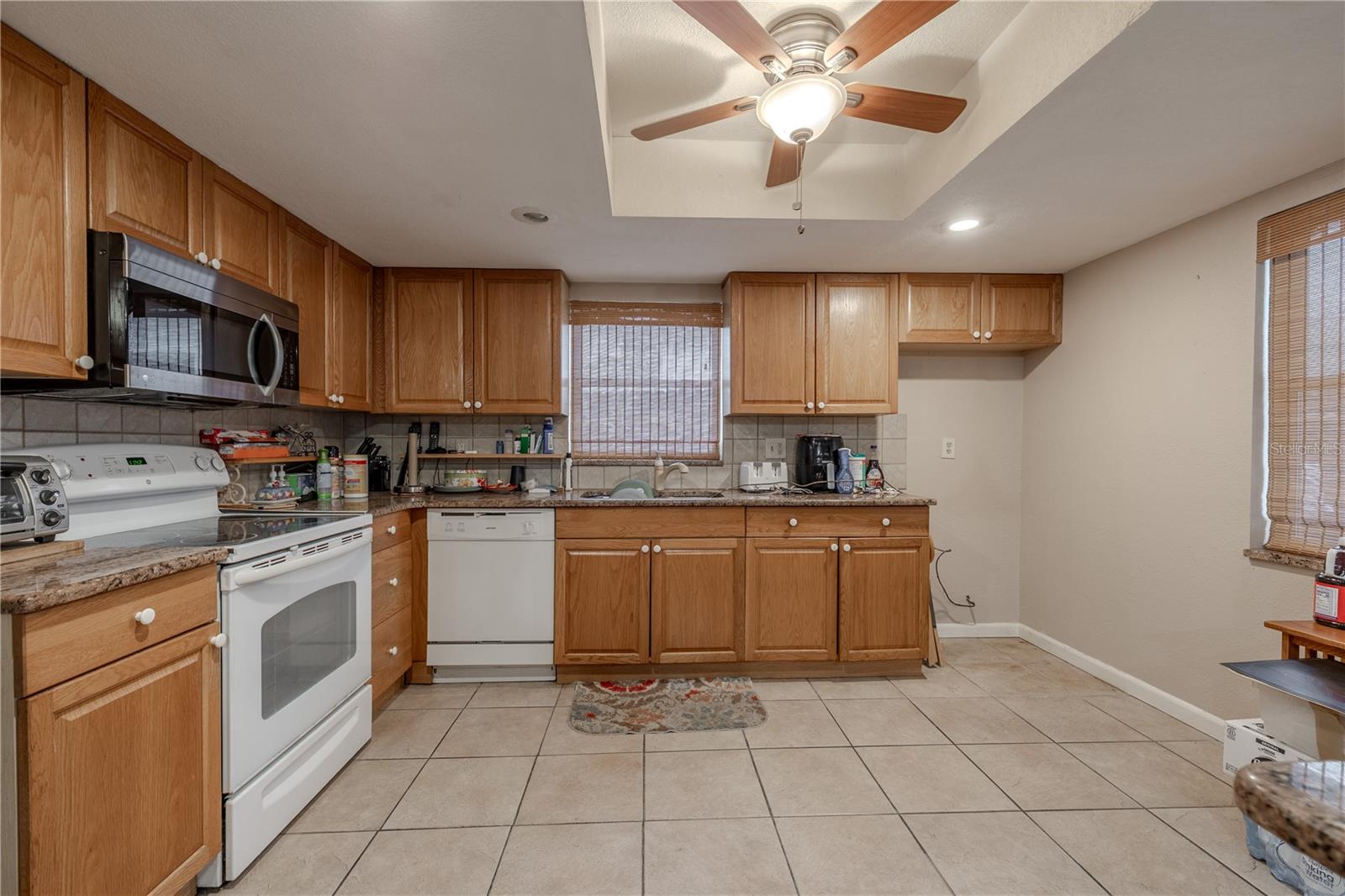 Bright and functional kitchen featuring abundant wood cabinetry, granite-look countertops, and a tile backsplash. A skylight with ceiling fan and a large window above the sink bring in plenty of natural light. The layout offers ample counter space for meal preparation, along with appliances that include range, microwave, and dishwasher.