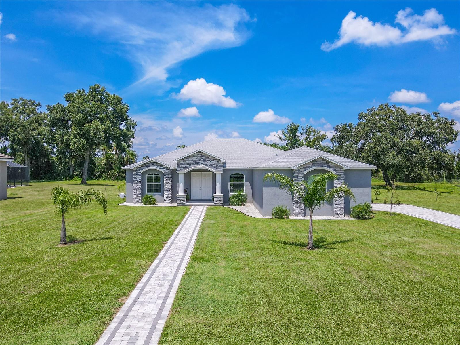 Set on a sprawling, well-manicured lot, this contemporary ranch-style home boasts a striking gray façade accented with elegant stonework and crisp white trim. A welcoming double-door entry is framed by stately columns and a covered front porch, while arched windows enhance the home’s curb appeal. The long paver walkway and matching driveway adds symmetry, leading the eye toward the home’s clean architectural lines.