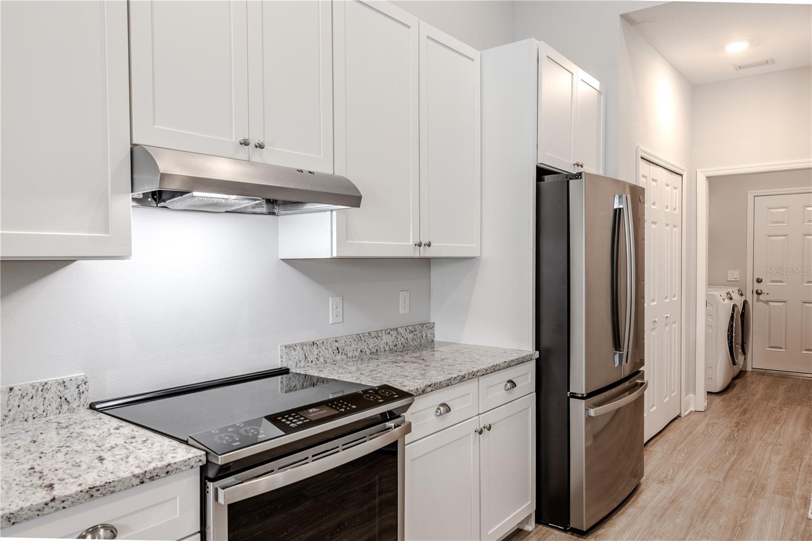 The kitchen features sleek stainless steel appliances, including a French door refrigerator and smooth-top range with a stainless vent hood. Crisp white cabinetry and granite countertops extend along both sides, providing ample prep and storage space. The layout offers convenient access to the laundry room and garage, making everyday tasks effortless.