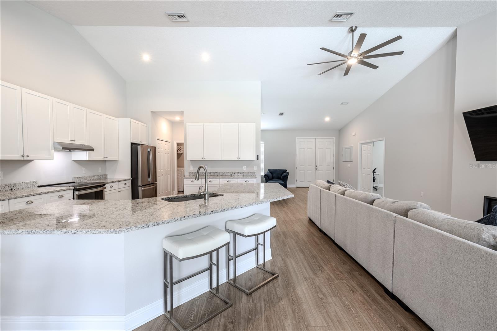 This open-concept kitchen and living area is designed for both style and function. The kitchen features an expansive granite island with an undermount sink, breakfast bar seating, and sleek white cabinetry complemented by stainless steel appliances.
