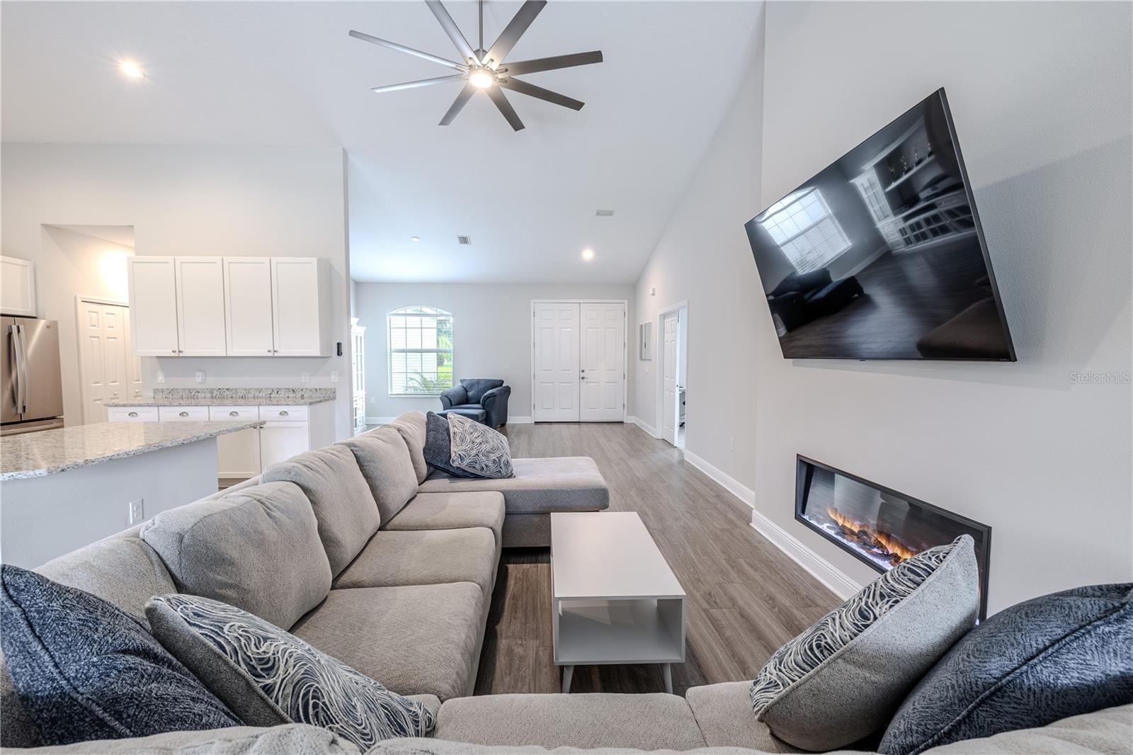 The family room offers a warm and inviting atmosphere, featuring a sleek electric fireplace creating a stylish focal point. High ceilings with a modern ceiling fan enhance the open feel, while wood-look flooring adds warmth and durability.