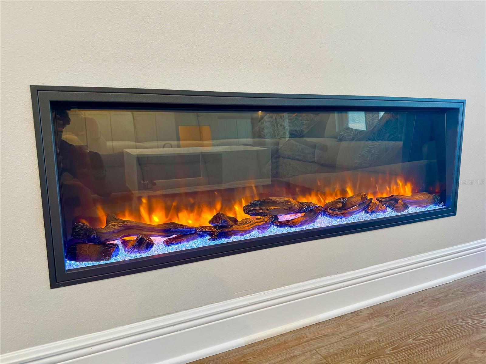 A sleek, wall-mounted electric fireplace serves as a stunning focal point in the family room, showcasing realistic flame effects with vibrant ember lighting over decorative logs. The modern black frame contrasts beautifully with the light wall color, adding both warmth and contemporary style to the space.