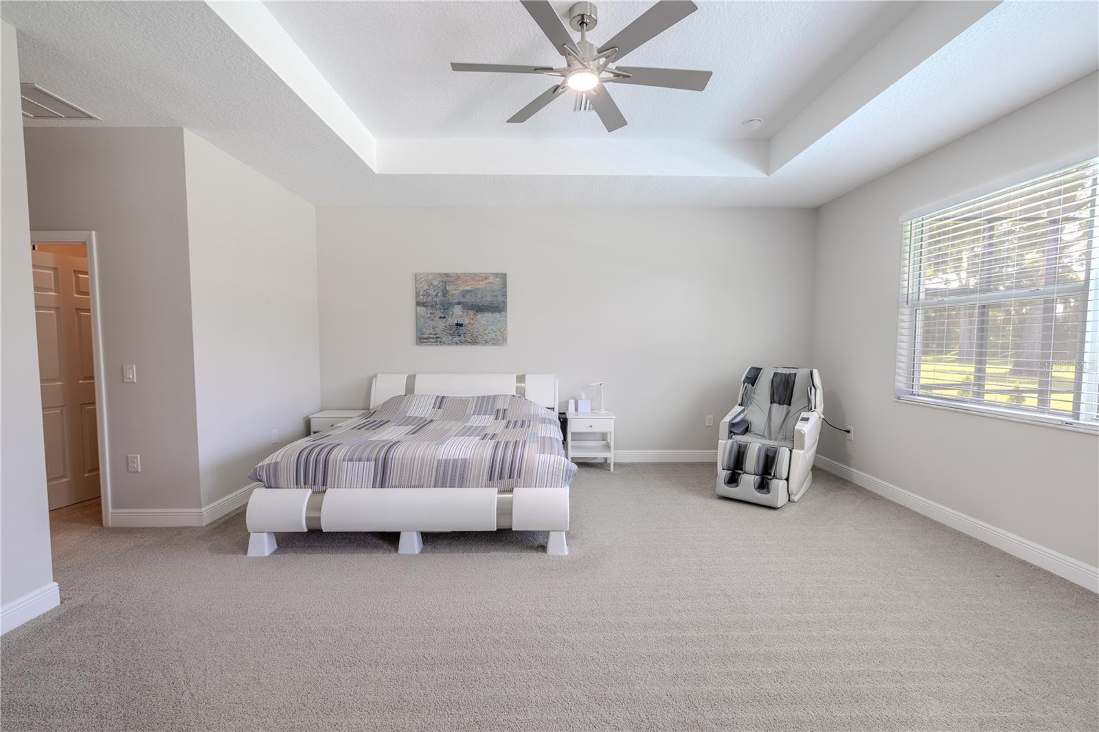 The primary bedroom features a stylish tray ceiling with recessed lighting and a modern ceiling fan, adding architectural interest. Plush carpeting and neutral tones and a large window fills the room with natural light. with views of the backyard.