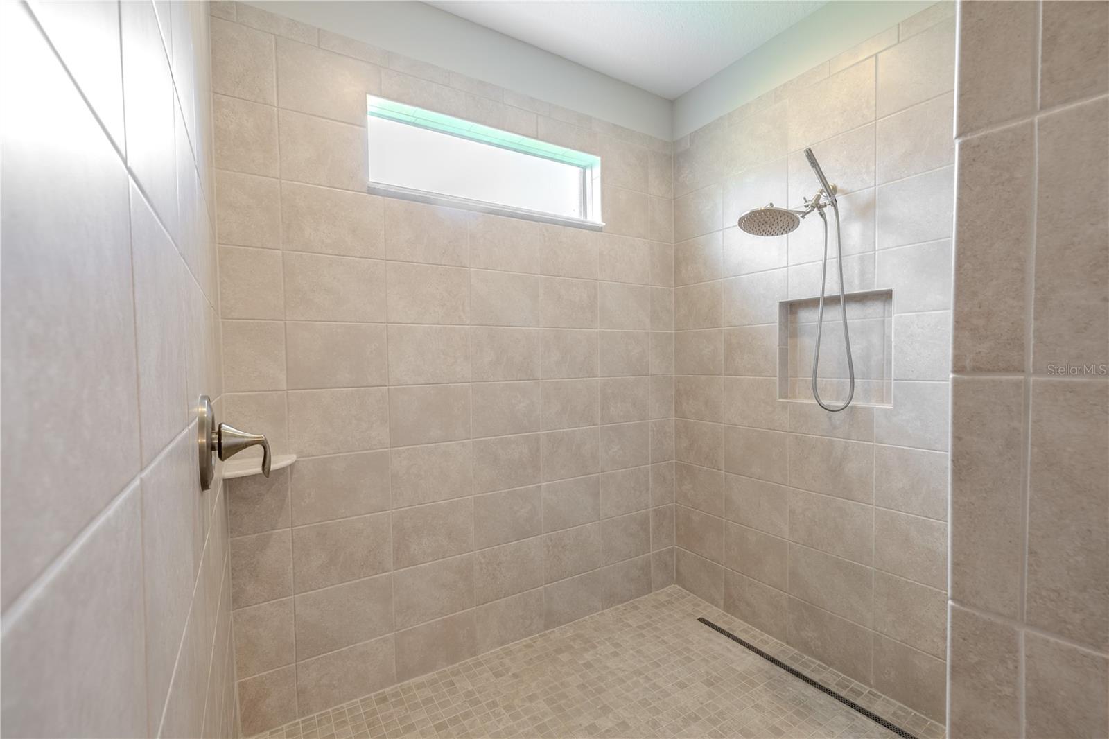 Primary Bathroom: The spacious walk-in shower showcases floor-to-ceiling neutral tile with a coordinating mosaic tile floor. A sleek frameless design features a built-in niche for toiletries, a horizontal transom window that brings in natural light while maintaining privacy, and a modern rainfall showerhead with handheld attachment for a spa-like experience.