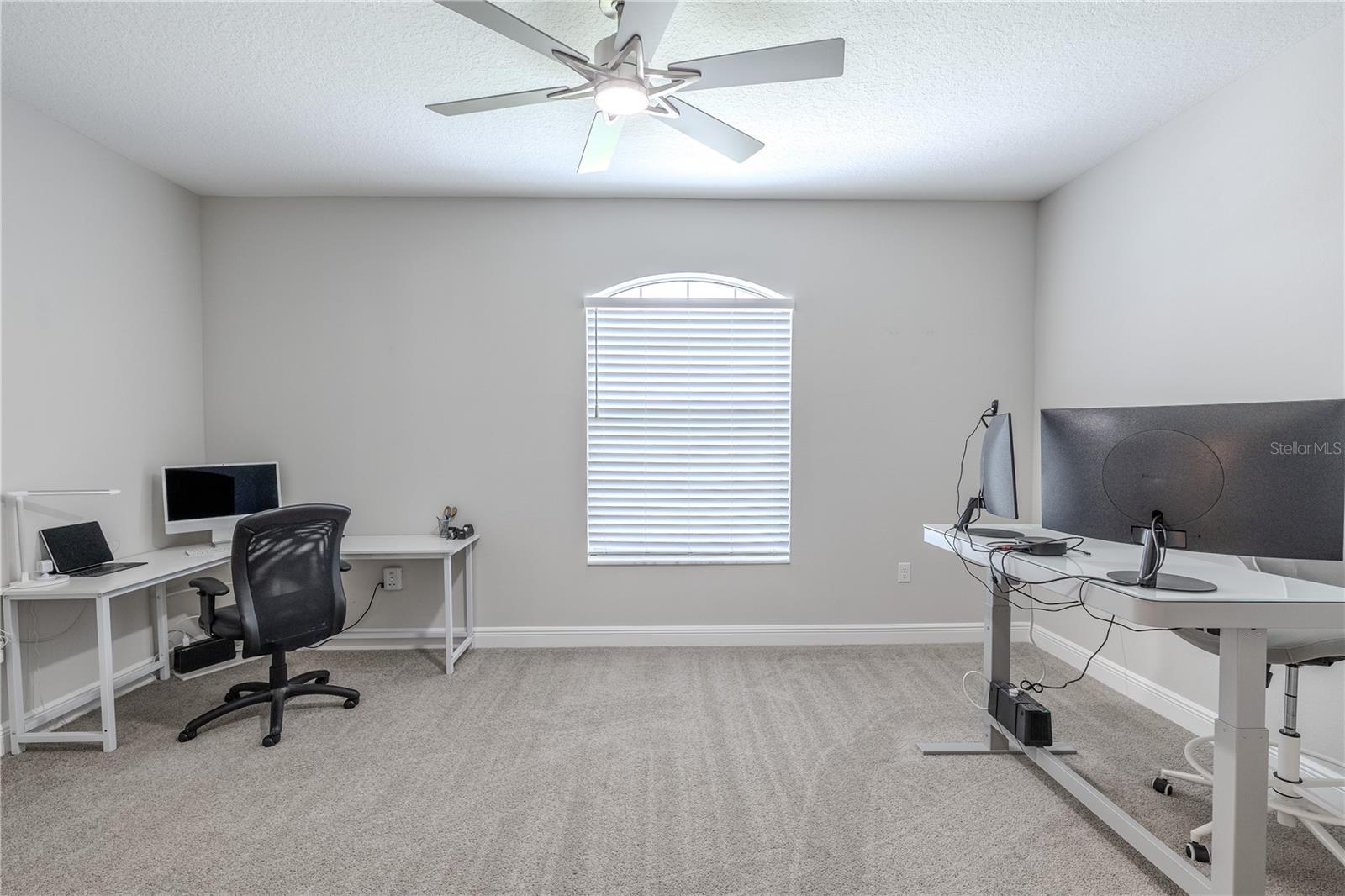 The office is a spacious, carpeted room with soft gray walls and a central ceiling fan with a light fixture. A large arched window with blinds allows for natural light when open.