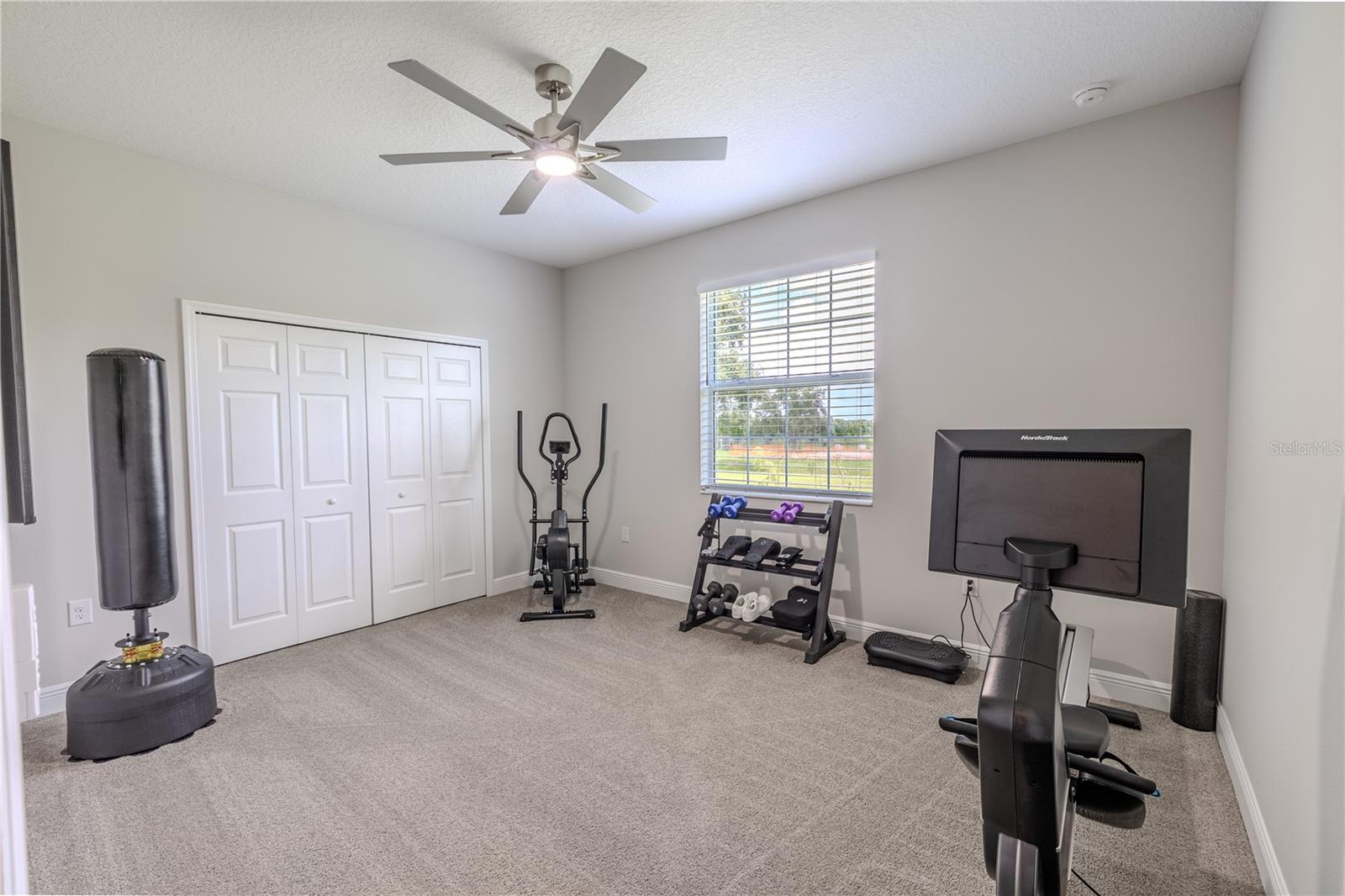 The second bedroom is a bright, carpeted space with neutral-toned walls and a central ceiling fan with a modern light fixture. A large window allows abundant natural light to flow in, offering pleasant views outside. Double white-paneled closet doors provide ample storage, while the open floor plan of the room offers versatility for various layouts or uses. The clean lines and uncluttered design create a fresh and inviting atmosphere.