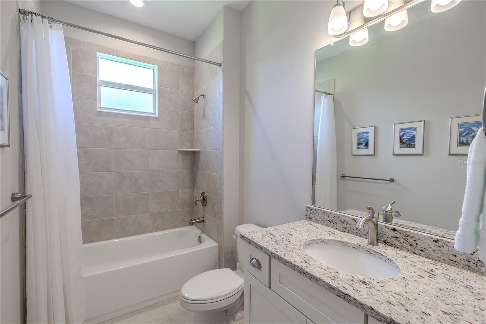 The second bathroom is bright and inviting, featuring a combination tub and shower with full-height neutral tile surround and a built-in corner shelf. A frosted window above the tub allows natural light while maintaining privacy. The single-sink vanity showcases a speckled granite countertop, under-mount sink, and brushed nickel faucet, paired with white cabinetry for a clean, modern look. Overhead, a large mirror is framed by a contemporary three-light fixture, enhancing the room’s open feel.