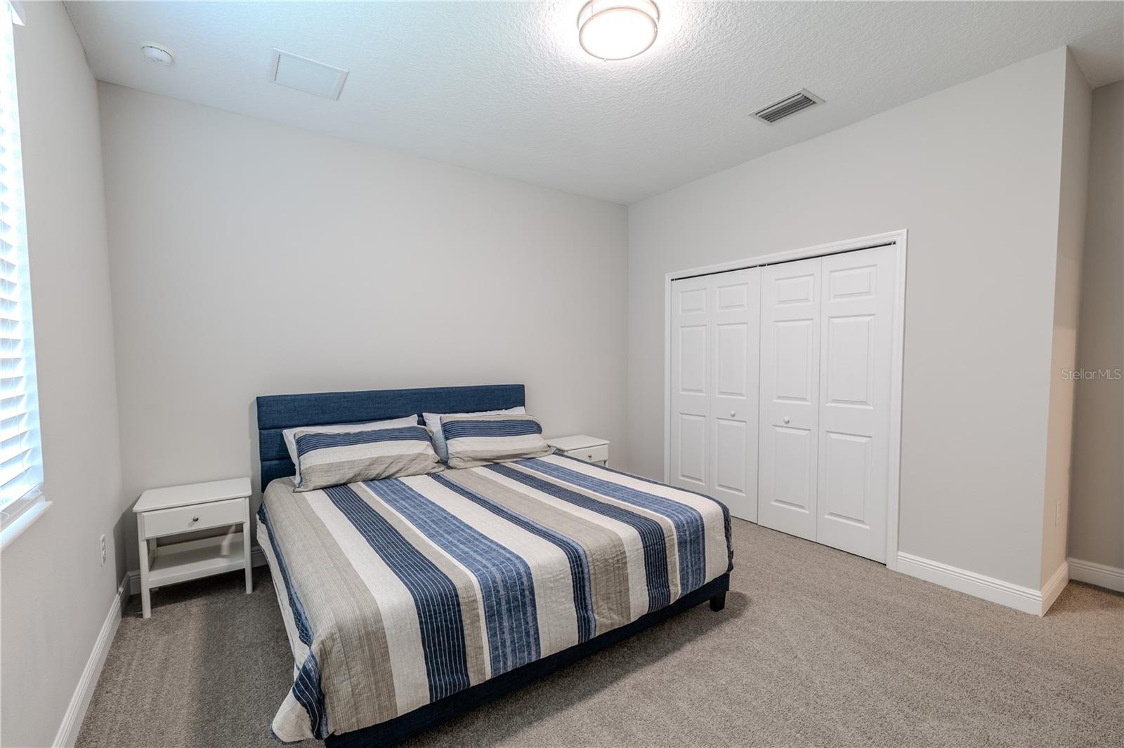 The third bedroom features soft, neutral carpeting and light-colored walls that create a bright, airy atmosphere. A flush-mounted ceiling light provides even illumination, while a window allows for natural light during the day. The room includes a double-door closet with white paneled doors, offering ample storage space. Clean lines and a simple design make it a versatile space for a variety of uses.