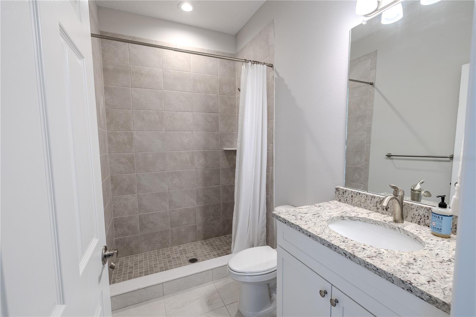 The third bathroom features a clean, modern design with a walk-in shower clad in neutral-toned tile, accented by a small built-in corner shelf for convenience. A white shower curtain adds a soft touch, complementing the light granite countertop that sits atop crisp white cabinetry. The undermount sink is paired with a brushed nickel faucet, matching the hardware throughout the space. A large frameless mirror above the vanity enhances the room’s brightness, while the light tile flooring ties the entire look together for a cohesive, fresh feel.