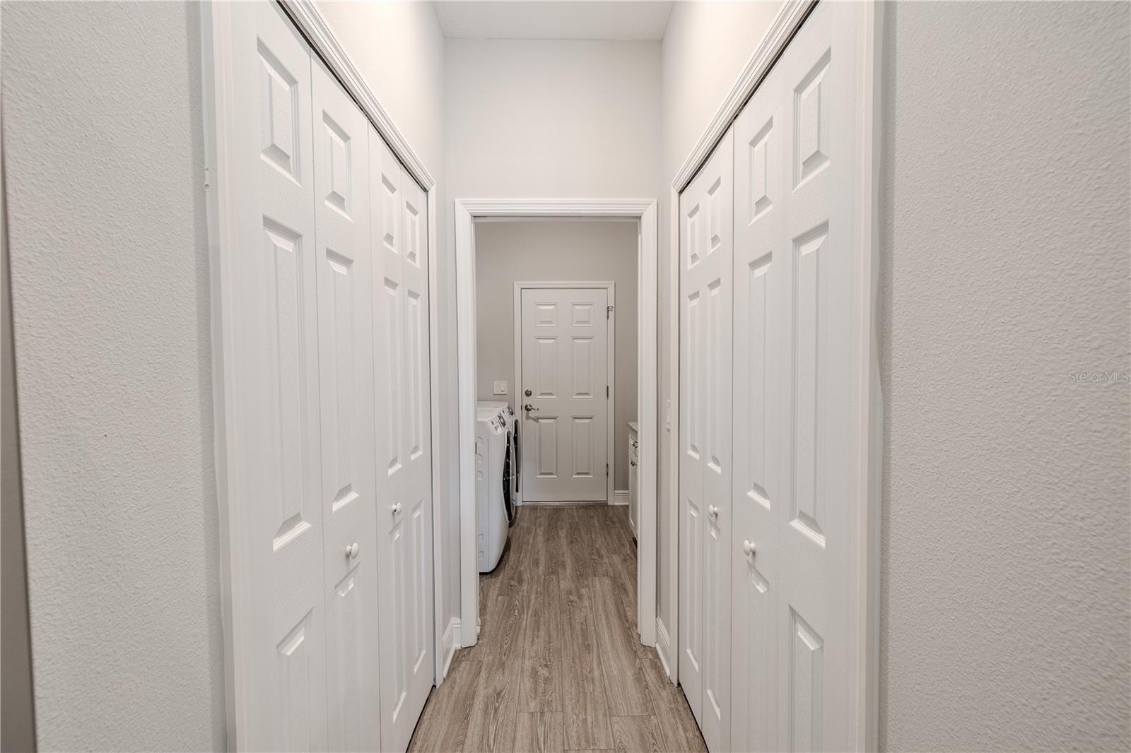 A hallway leads to the laundry room which is bright and functional, featuring a clean, neutral palette with light-colored walls and wood-look flooring that adds warmth and texture. On either side, there are multiple sets of double doors with white paneled finishes, providing ample closet and storage space. The streamlined layout creates an efficient transition from the main living areas to the laundry space.